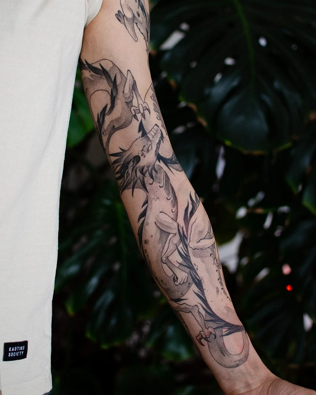 A person's arm with a large black and gray tattoo of a dragon and a figure of a person with long hair and a tail, set against a dark leafy background.