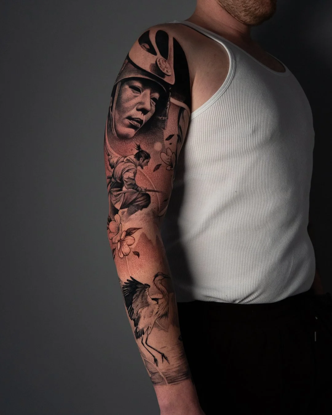 Close-up of a person's arm tattoo featuring a woman's face, a seated warrior with a sword, flowers, and a heron, against a dark background.