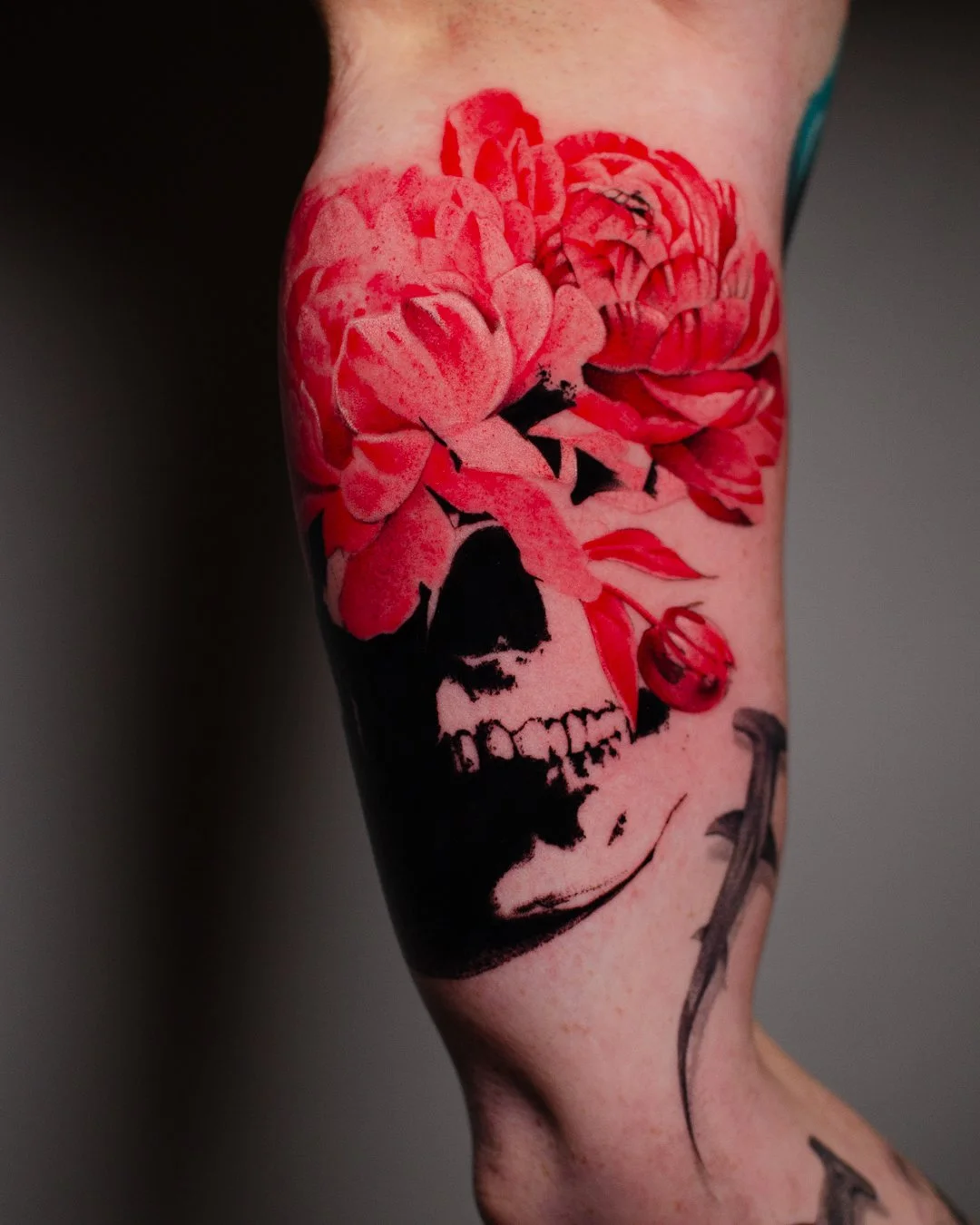 Tattoo of a skull with large pink peonies and a small red flower on a person's arm.