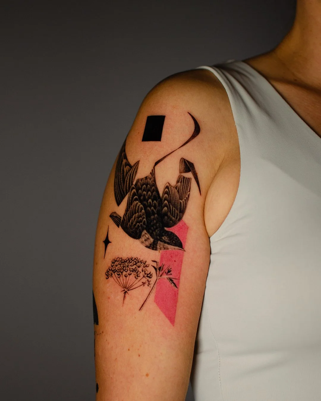 Close-up of a person's upper arm tattoo featuring a bird, geometric shapes, and floral elements, with a sleeveless white shirt.