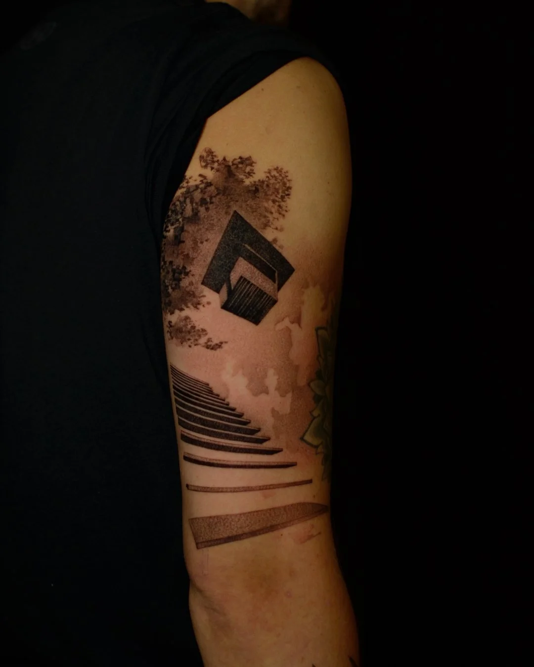 Tattoo of stairs leading to a floating house with trees and clouds in a monochrome style on a person's upper arm.
