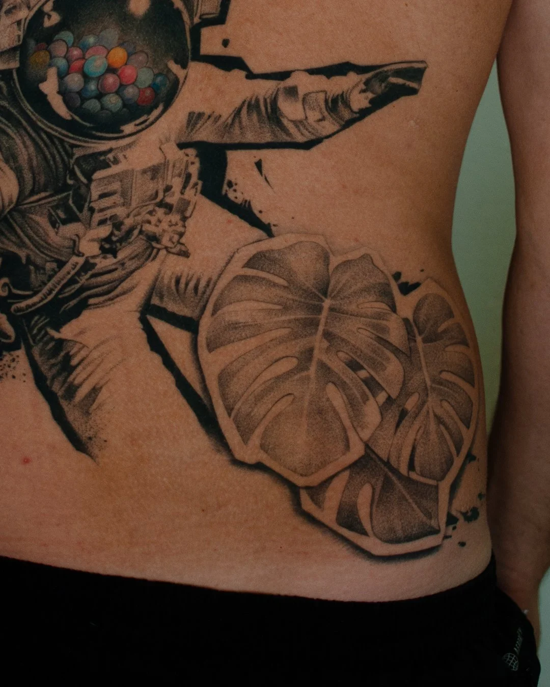 Tattoo of a vintage space helmet with colorful balls inside and a detailed Monstera leaf on the person's side.