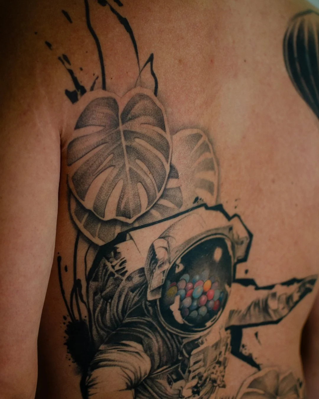 Tattoo of a monstera leaf and an astronaut helmet with colorful balls inside, on a person's chest.