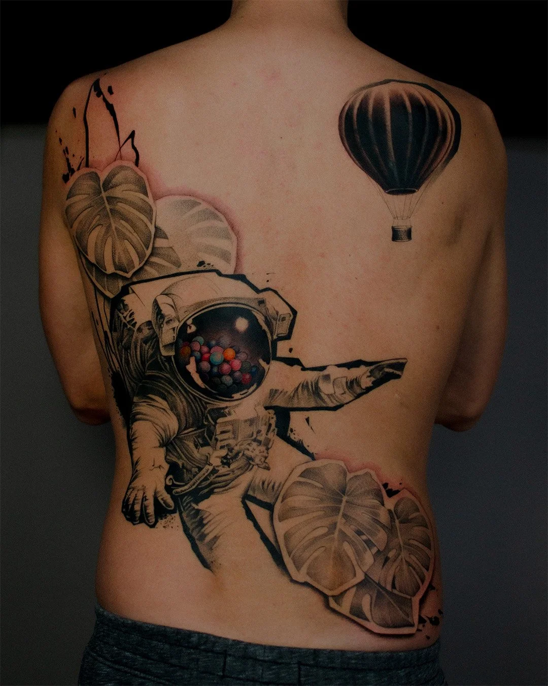 Tattoo of an astronaut floating among large leaves with a hot air balloon in the background on a person's back.