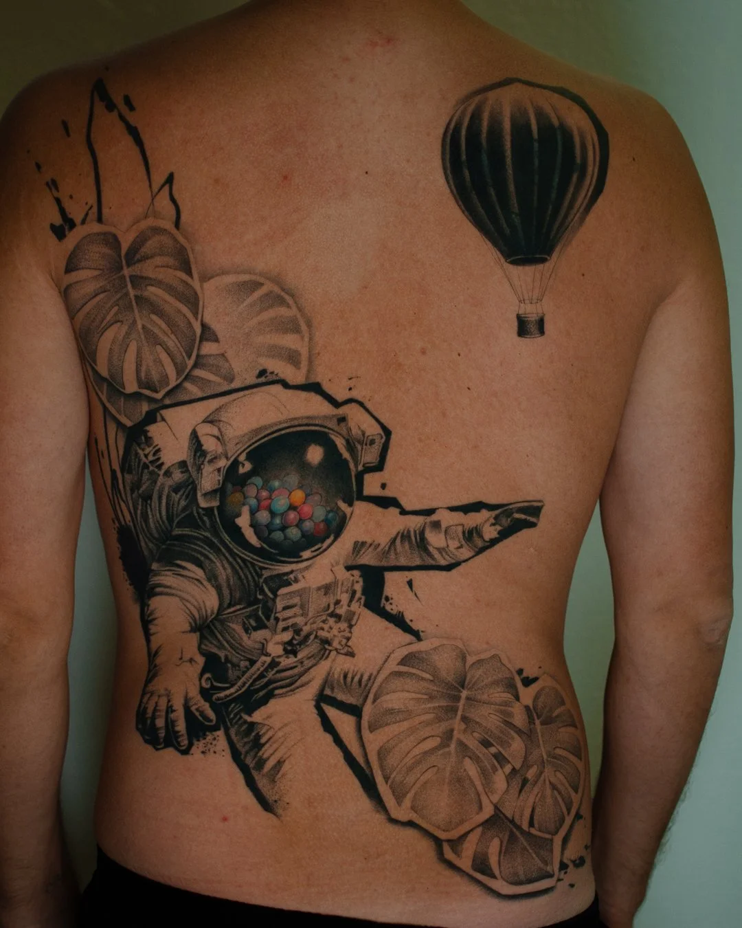 Tattoo of an astronaut pointing, surrounded by tropical leaves and a hot air balloon on the chest.