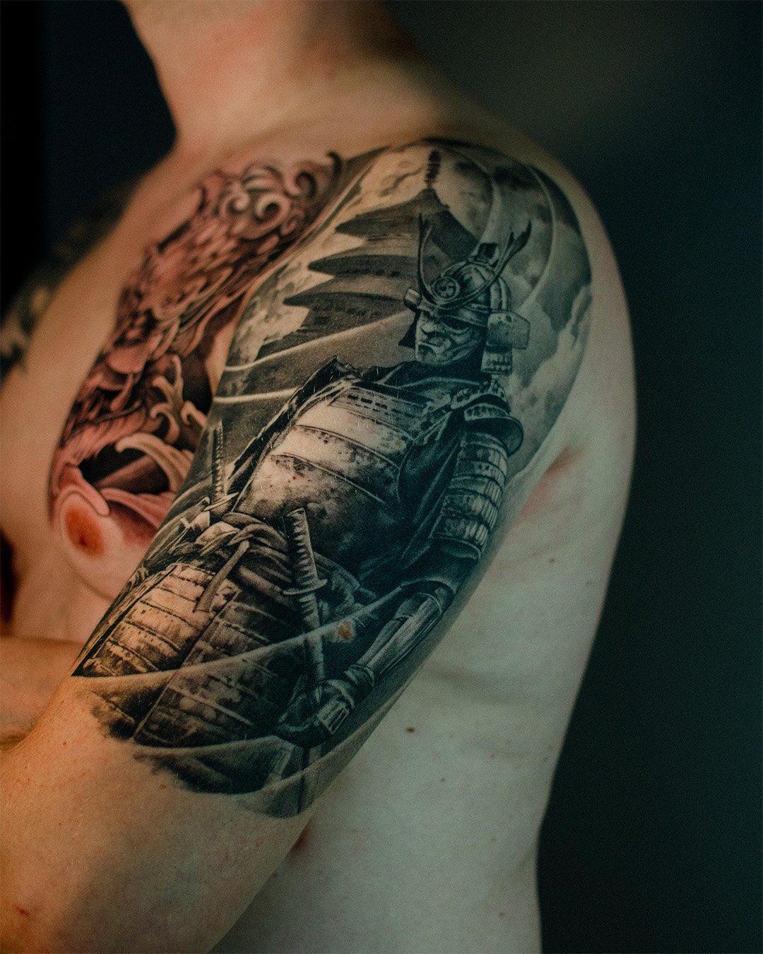 Black and gray tattoo of a samurai warrior in armor on the upper arm, with traditional Japanese elements in the background.