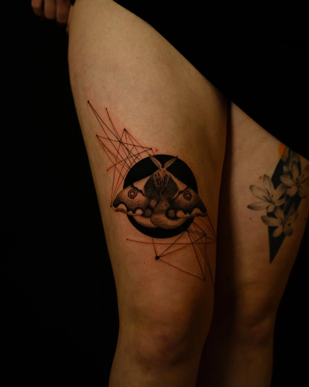 Tattoo of a moth within a black circle, with geometric lines extending outward on a person's upper thigh.