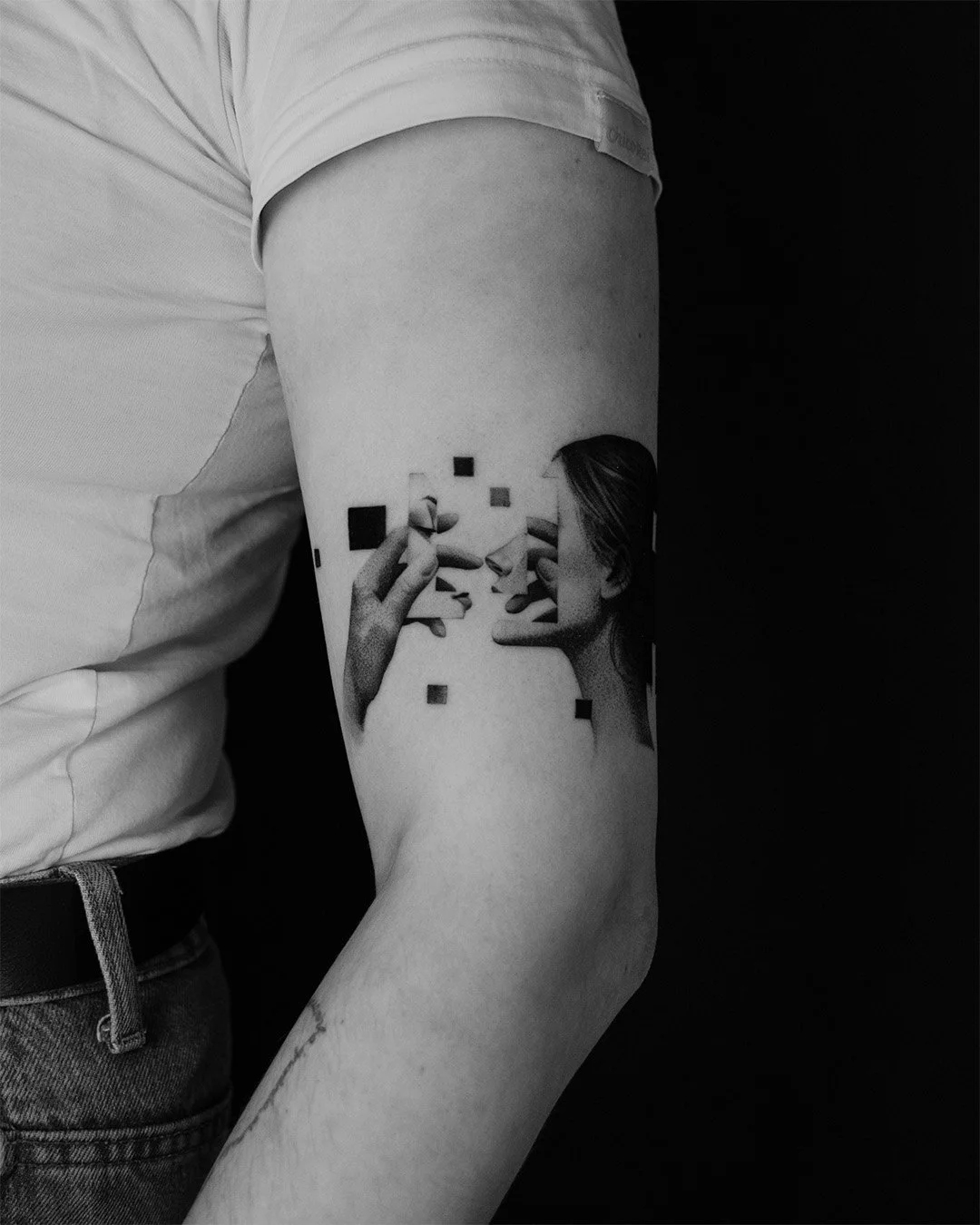 Black and white tattoo of two women facing each other, each holding a pen, creating a surreal image with geometrical shapes and the women appearing to draw or touch each other.