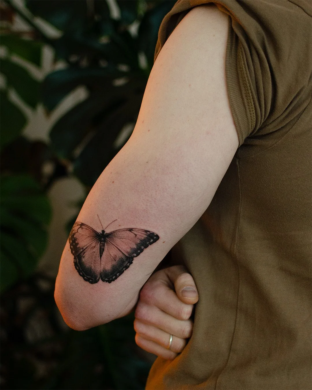 A person's upper arm with a butterfly tattoo, wearing a brown short-sleeved shirt, against a background of green leaves.