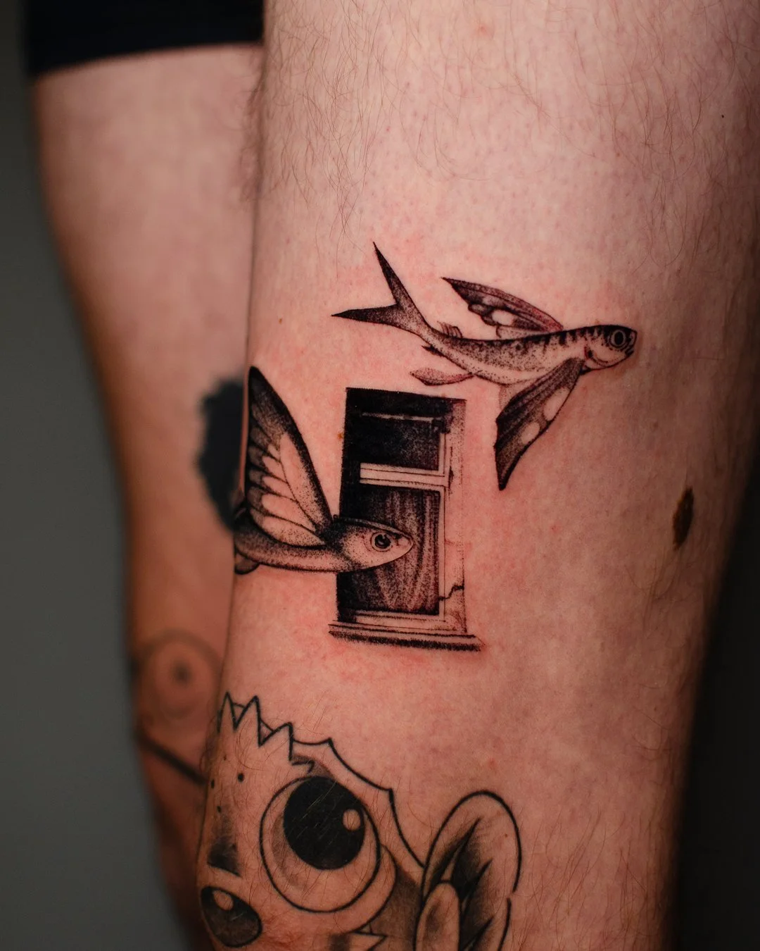Tattoo of two flying fish near a small window with open shutters on skin.