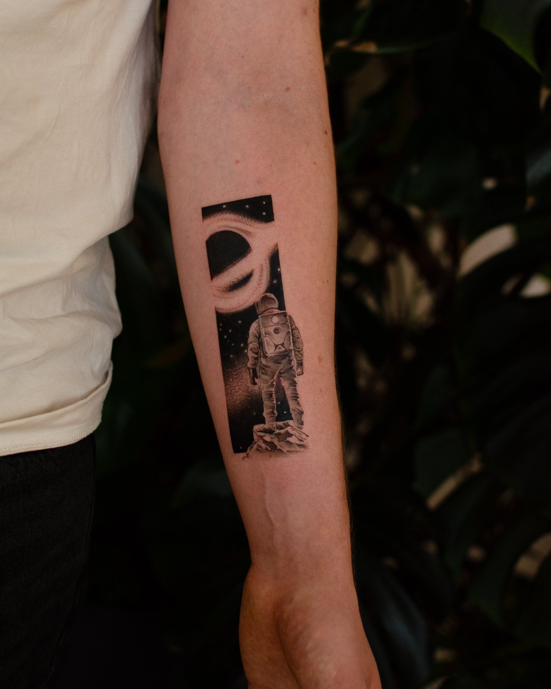Tattoo of an astronaut standing on a rock, looking at a galaxy and a planet in space, on a person's forearm.