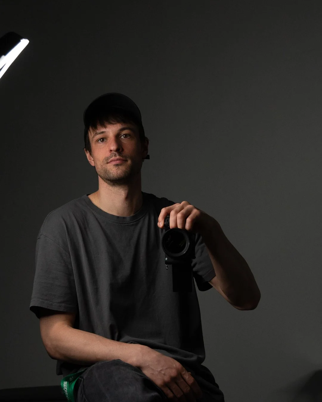 Tattoo artist, A young man with dark hair, wearing a black baseball cap and a dark gray T-shirt, taking a mirror selfie with a camera in a dimly lit room with a gray background.