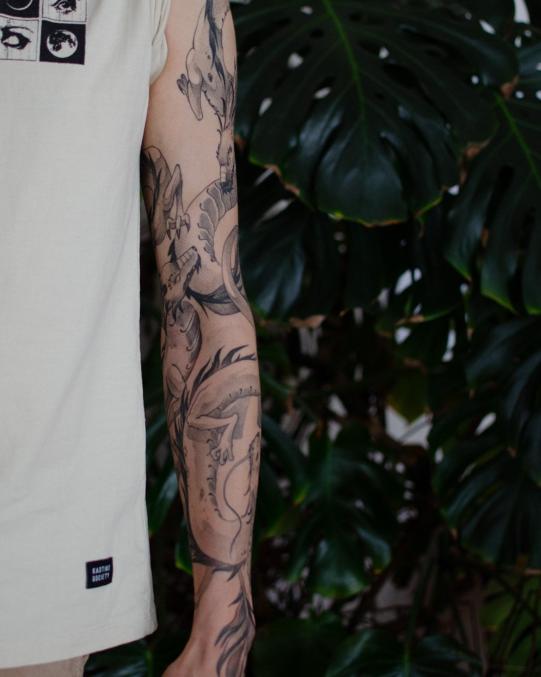 Close-up of a person's arm with a black and gray tattoo of a dragon, standing in front of large green leaves.