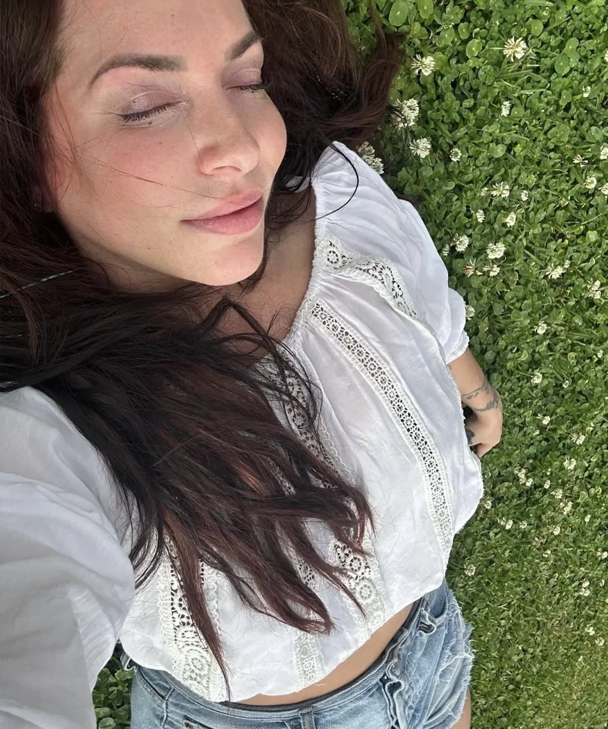 Woman with long dark hair lying on grass with small white flowers, eyes closed, wearing a white top with lace details and denim shorts.