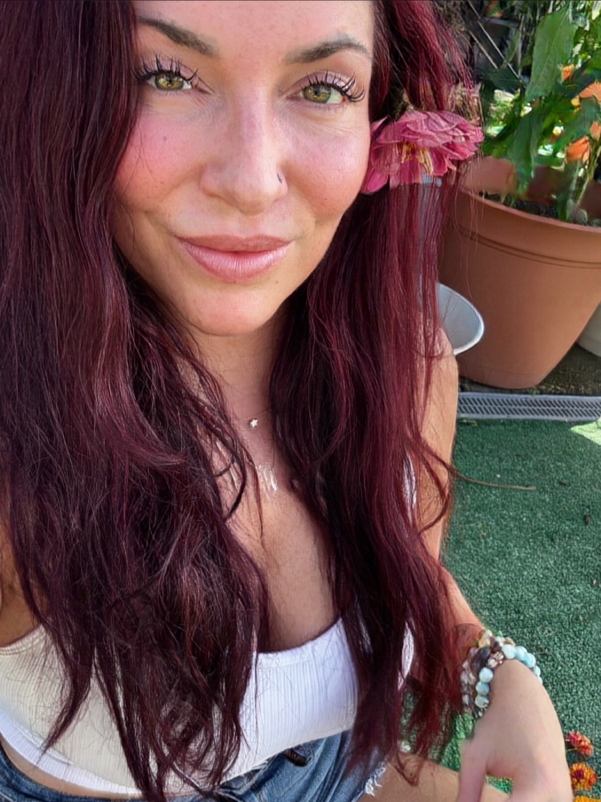 A woman with long red hair, green eyes, and a nose piercing, smiling outdoors near potted plants and flowers.