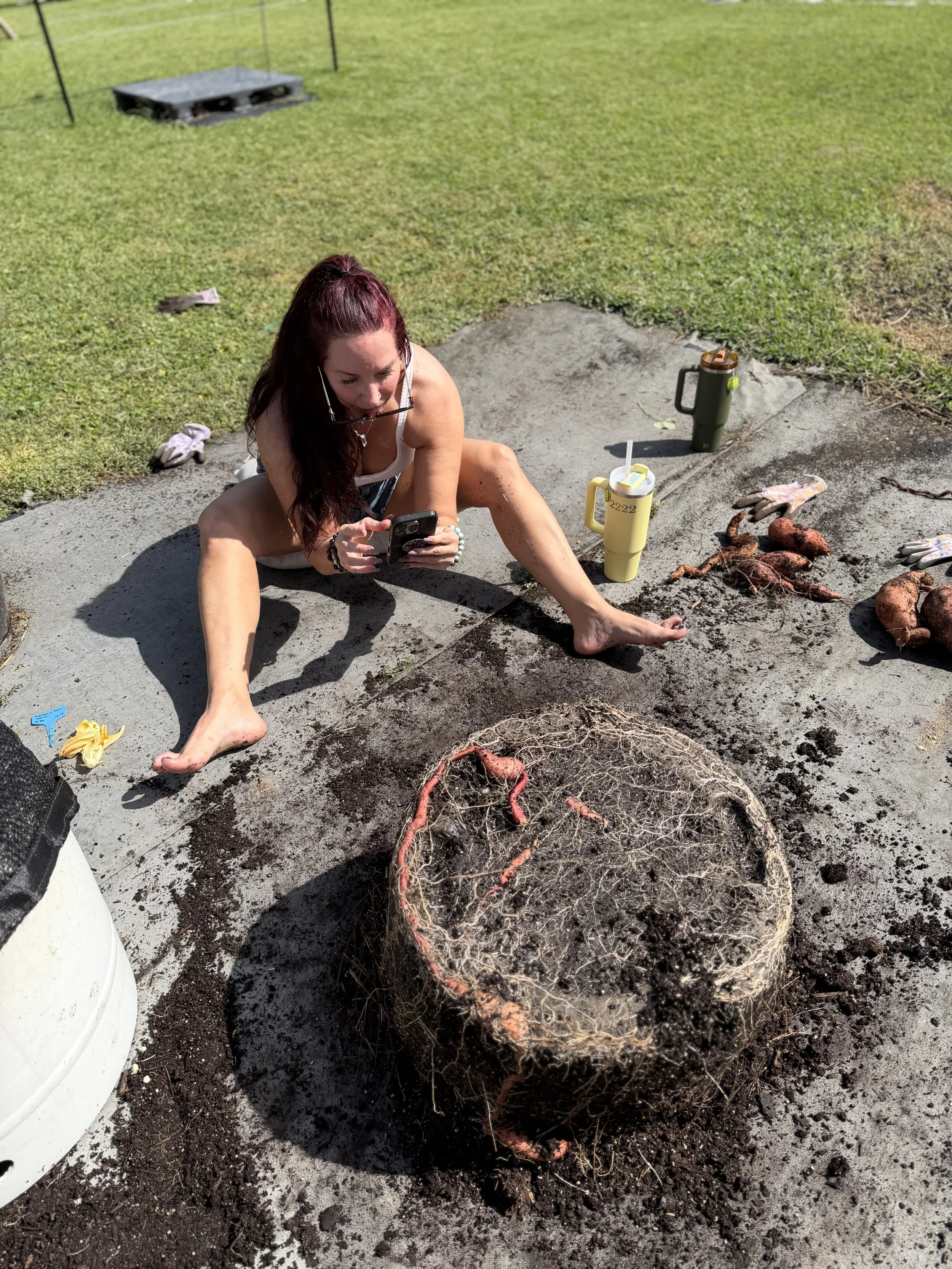 Woman sitting on the ground outdoors among clay and roots, looking at her phone, with gardening tools and plants nearby.