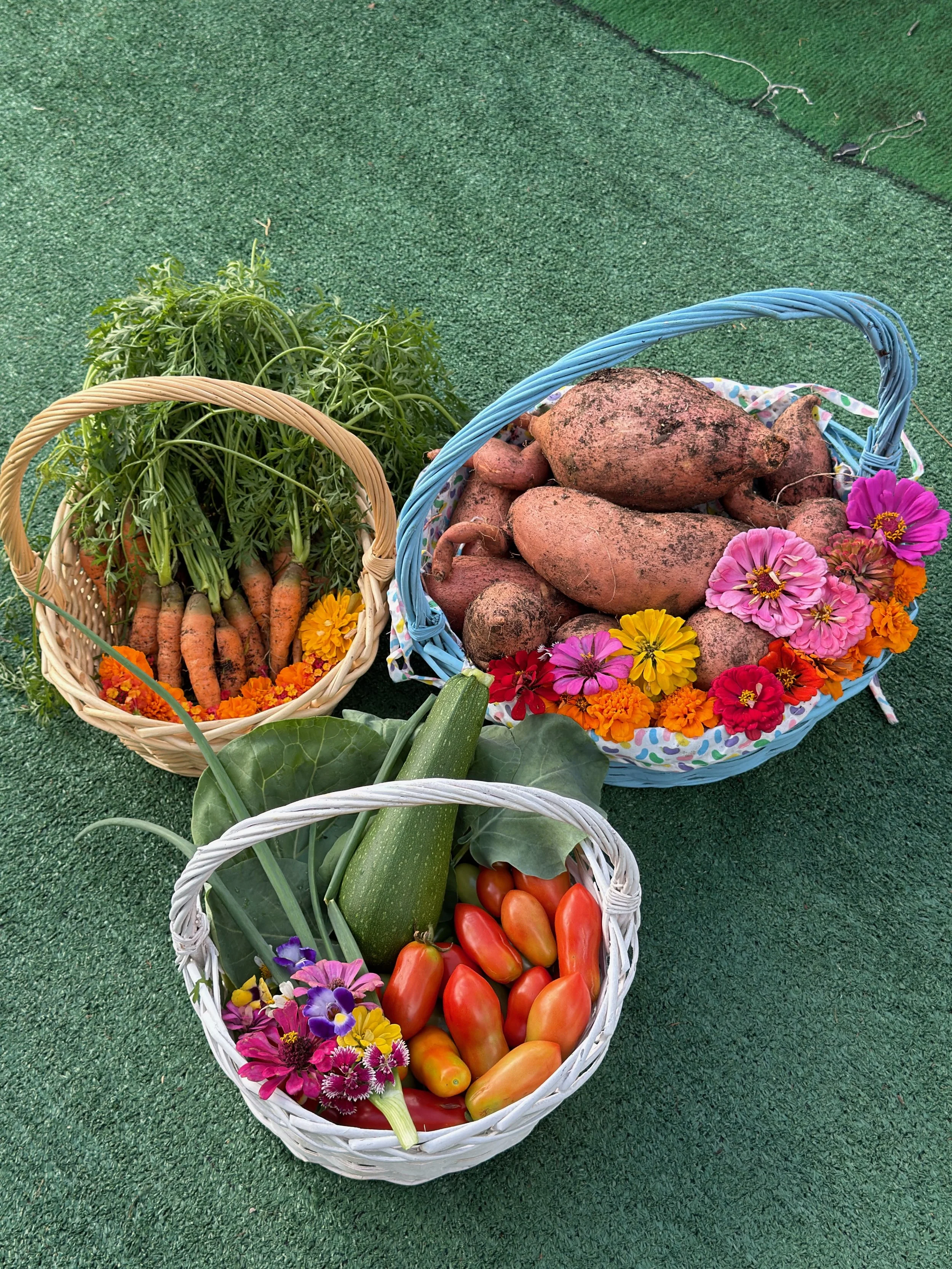 Three baskets of fresh vegetables on a green surface. The left basket contains carrots with green tops and decorated with marigold flowers. The middle basket holds dirt-covered sweet potatoes, decorated with pink, orange, and red flowers. The front basket has cherry tomatoes, a zucchini, green onions, and a small bouquet of pink, purple, and yellow flowers.