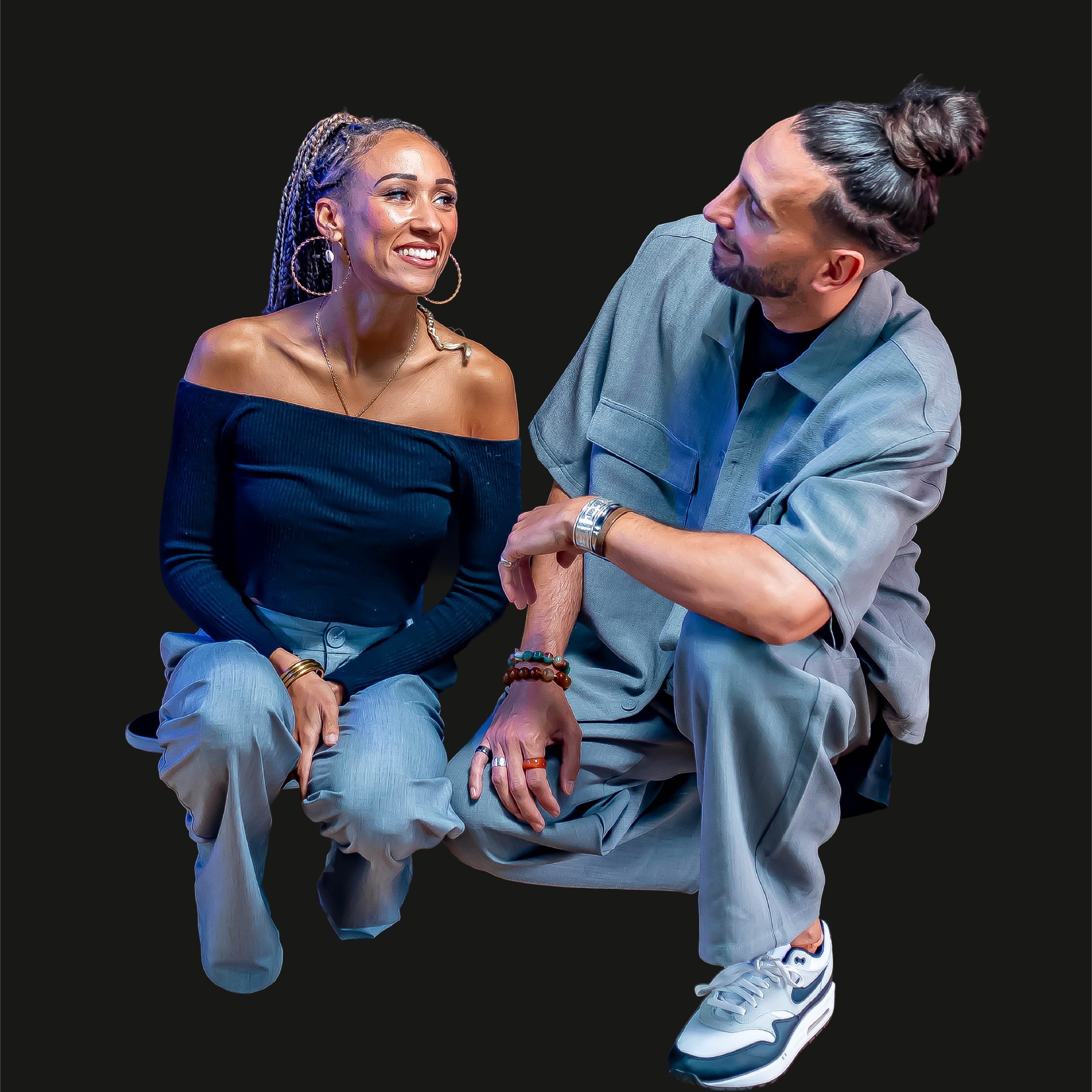 Deux personnes assises l'une à côté de l'autre contre un fond noir. La femme porte un haut noir, des jeans clairs et des accessoires en or, avec des cheveux en tresses bleues. L'homme porte une chemise grise, des jeans, et des baskets. Il a des cheveux en chignon et porte des bracelets colorés.