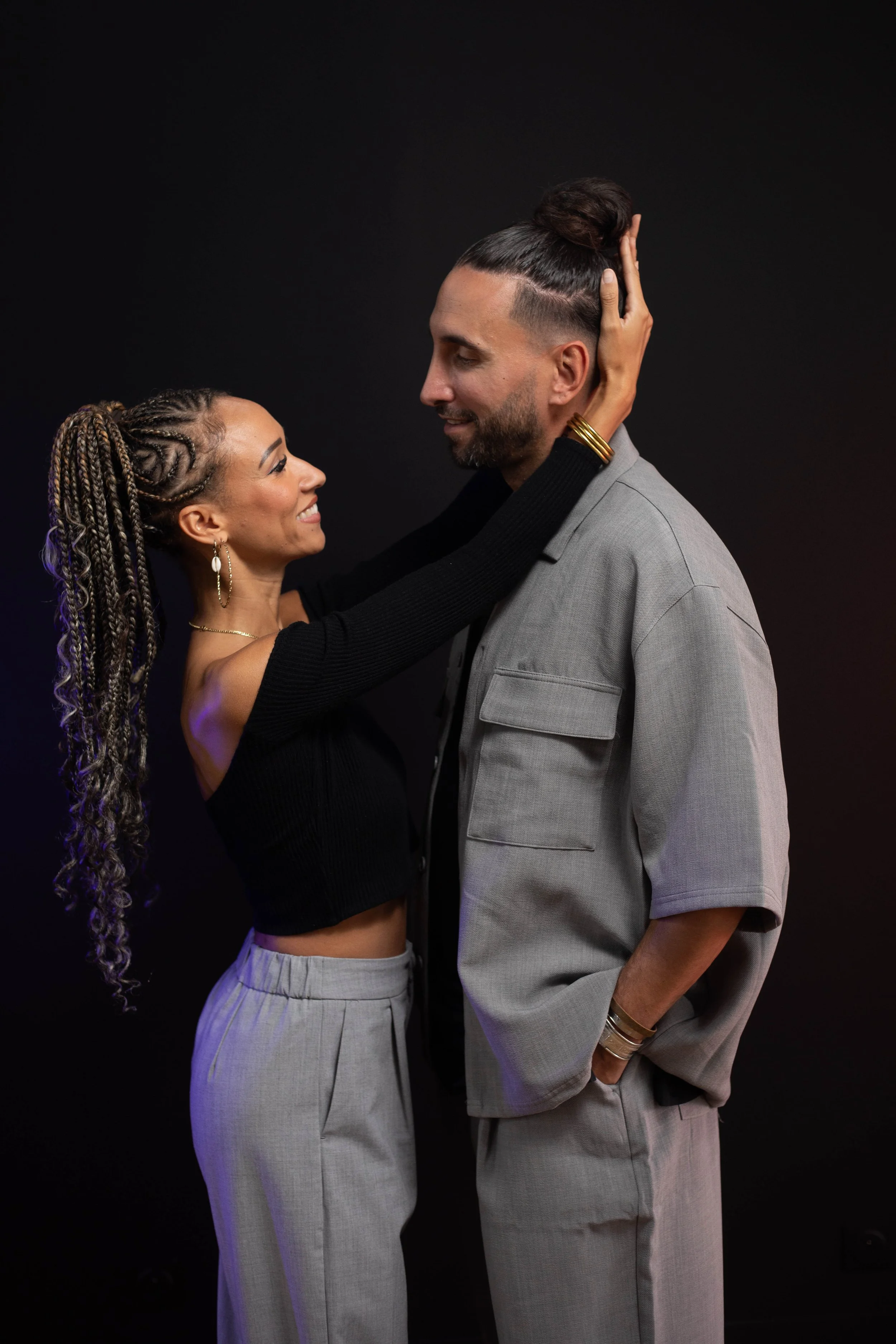 Un homme et une femme se regardent, la femme caresse la tête de l'homme, ils sourient. La femme a des cheveux en tresses et porte des boucles d'oreilles, l'homme porte un costume gris.