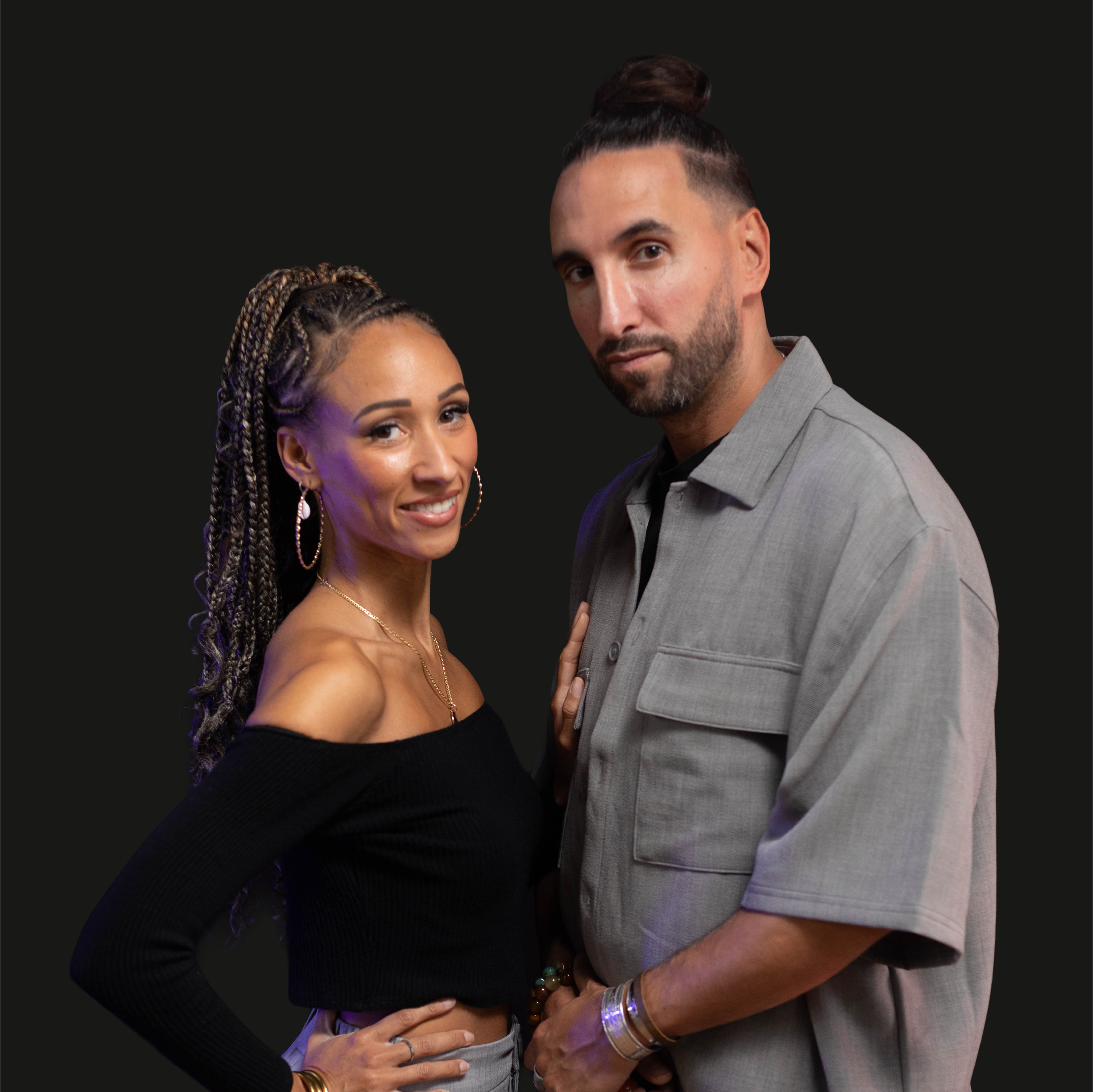Deux personnes, un homme et une femme, posant côte à côte contre un fond noir. La femme a des cheveux en tresses blondes et portait un haut noir. L'homme a une barbe, porte une veste gris clair et une chemise noire. Ils sont proches l'un de l'autre.