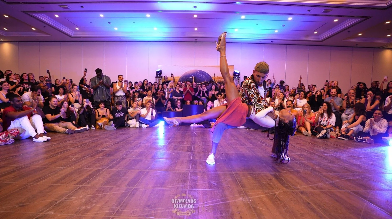 Deux danseurs de kizomba exécutent une acrobatie sur une piste en bois, entourés d'un public nombreux assis et debout, lors d'un spectacle ou compétition, avec éclairage coloré.