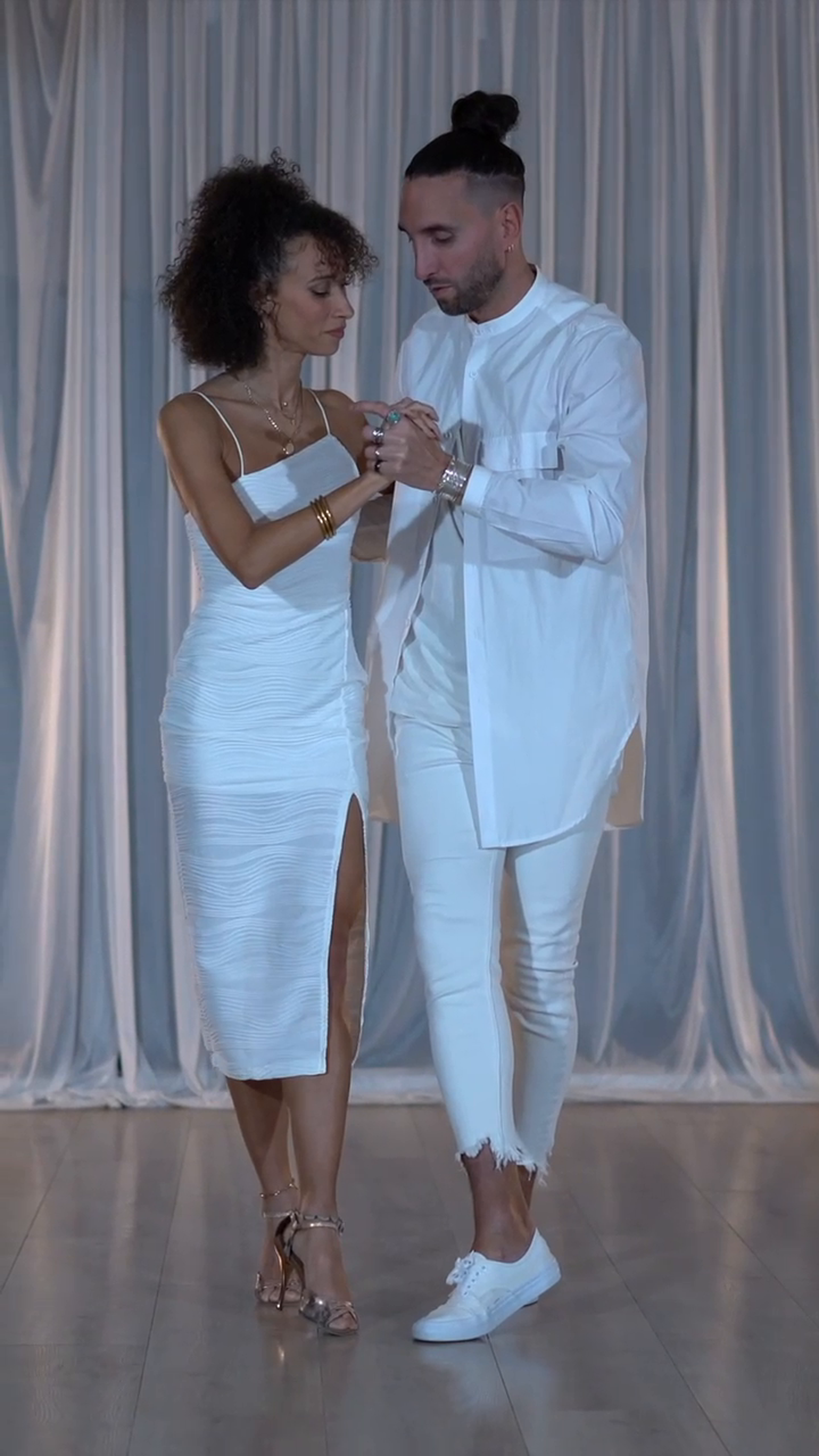 Un couple danse sur scène avec un fond de rideau blanc, tous deux habillés en blanc, la femme porte une robe longue avec une fente et des chaussures à talons, l'homme porte un chemisier et un pantalon déchiré à la cheville.