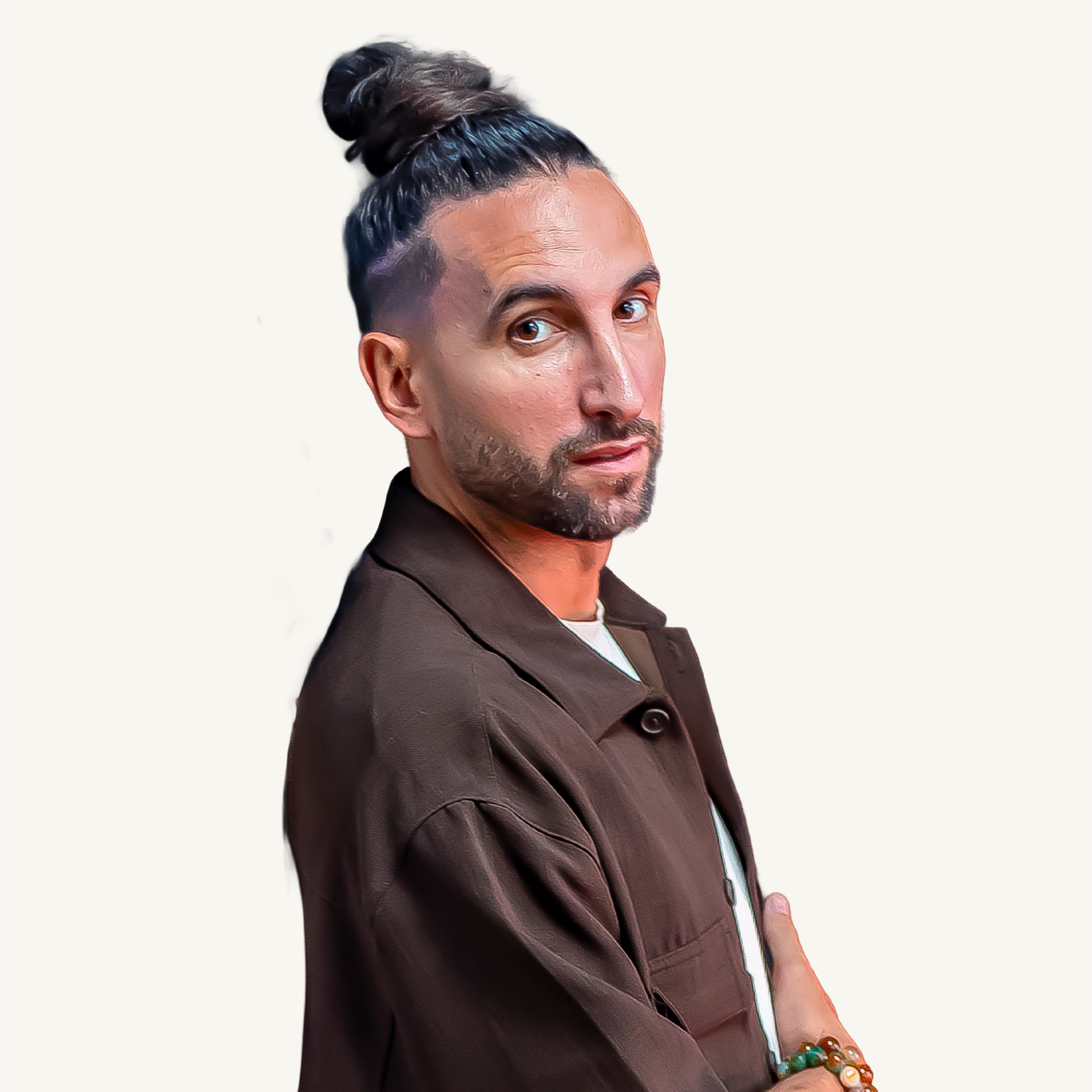 Un homme avec une barbe courte, portant une veste marron avec une chemise blanche, regarde vers la caméra, portant un bracelet coloré au poignet droit.