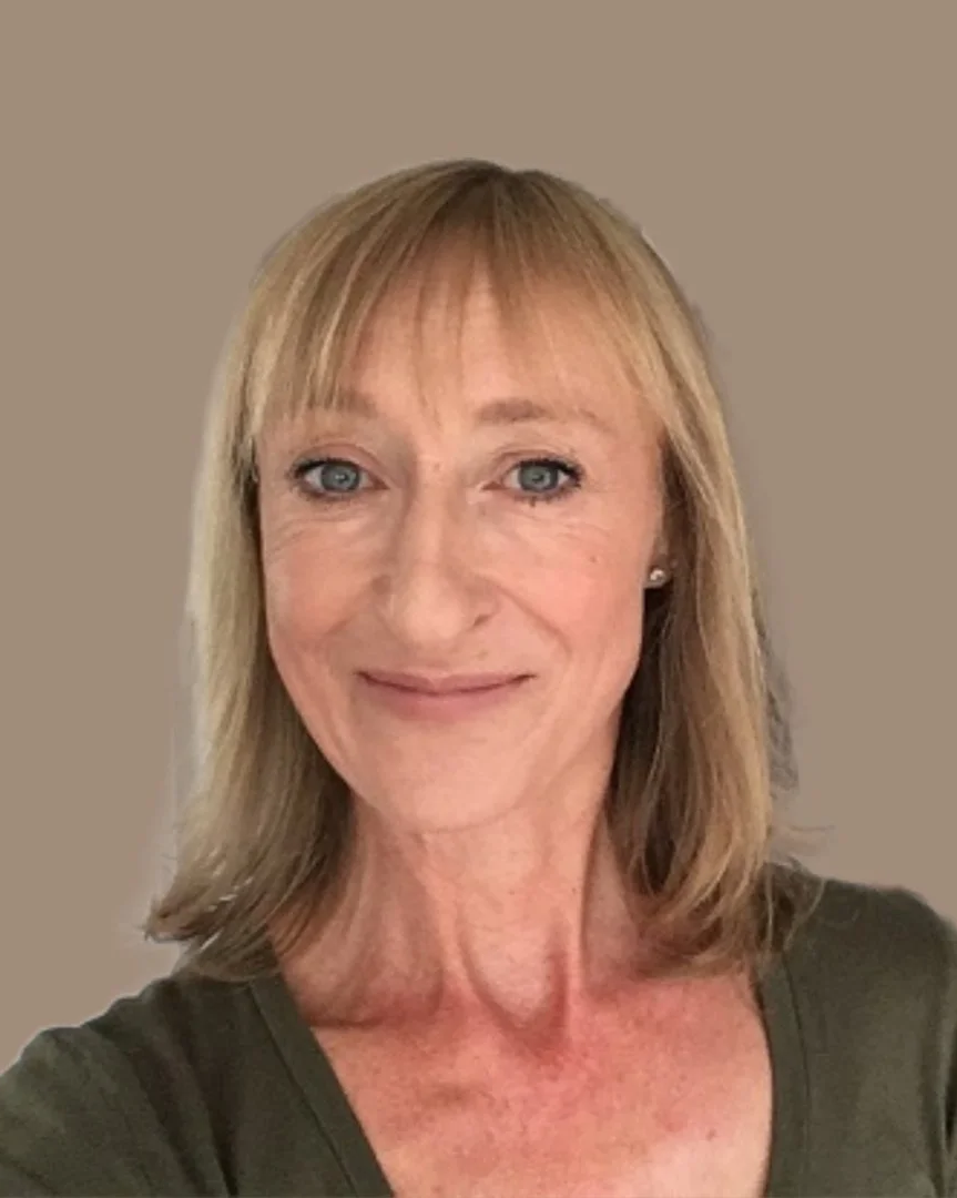A middle-aged woman with blonde hair and blue eyes smiling at the camera, wearing a dark green top, with a plain neutral-colored background.