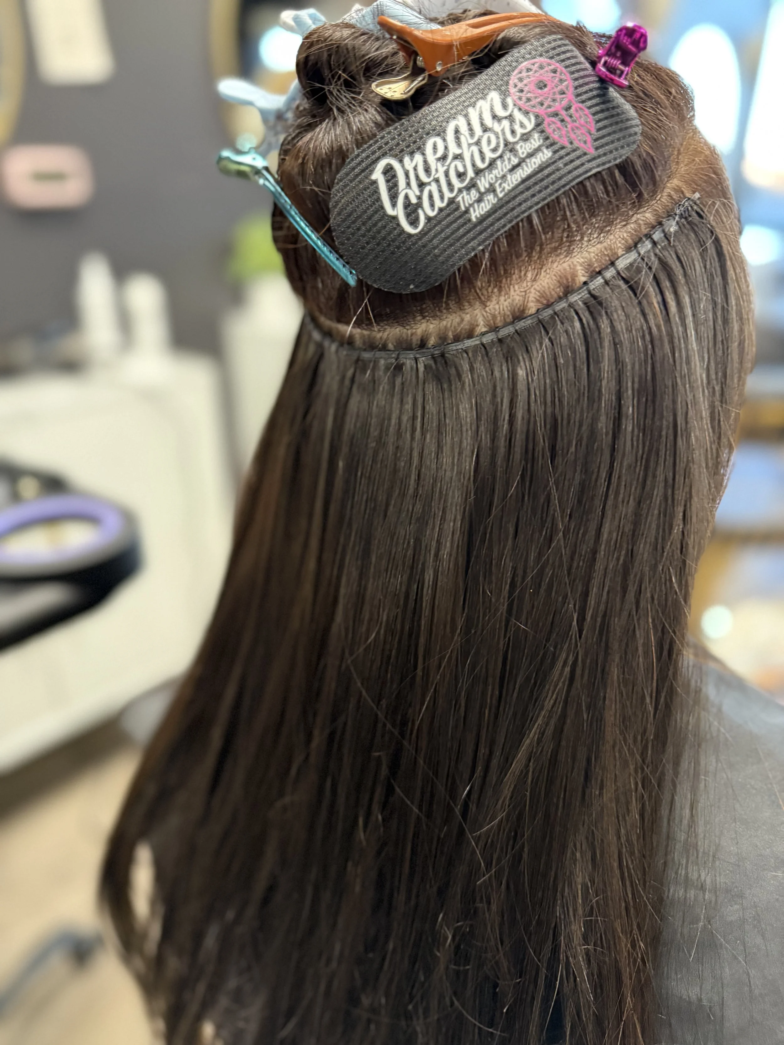 A person at a hair salon undergoing hair extensions, with hair clips and a headband labeled "Dream Catchers" holding hair in place.