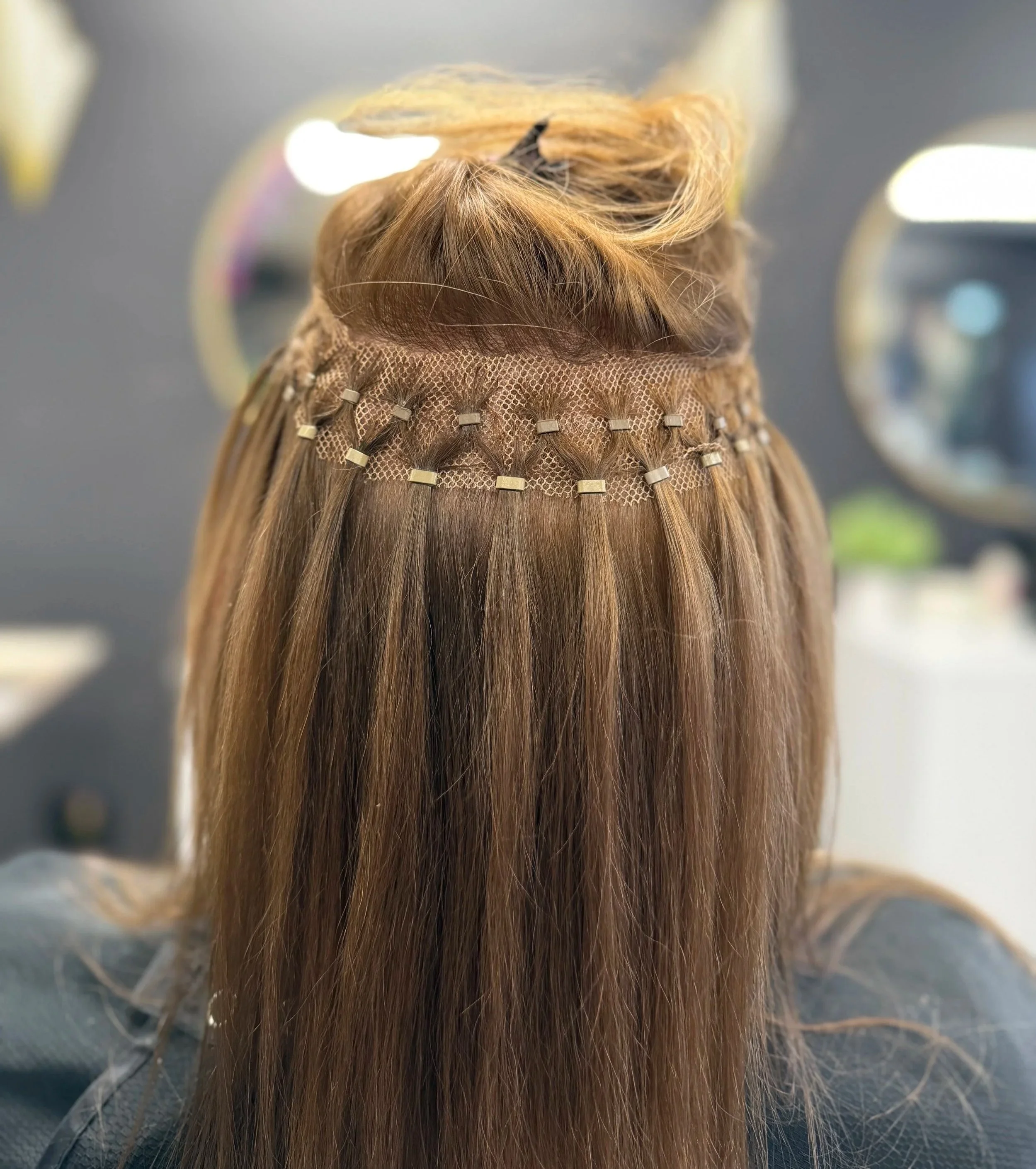 Back of a person with long brown hair, wearing a hair extension net and clips, in a salon setting with round mirrors.