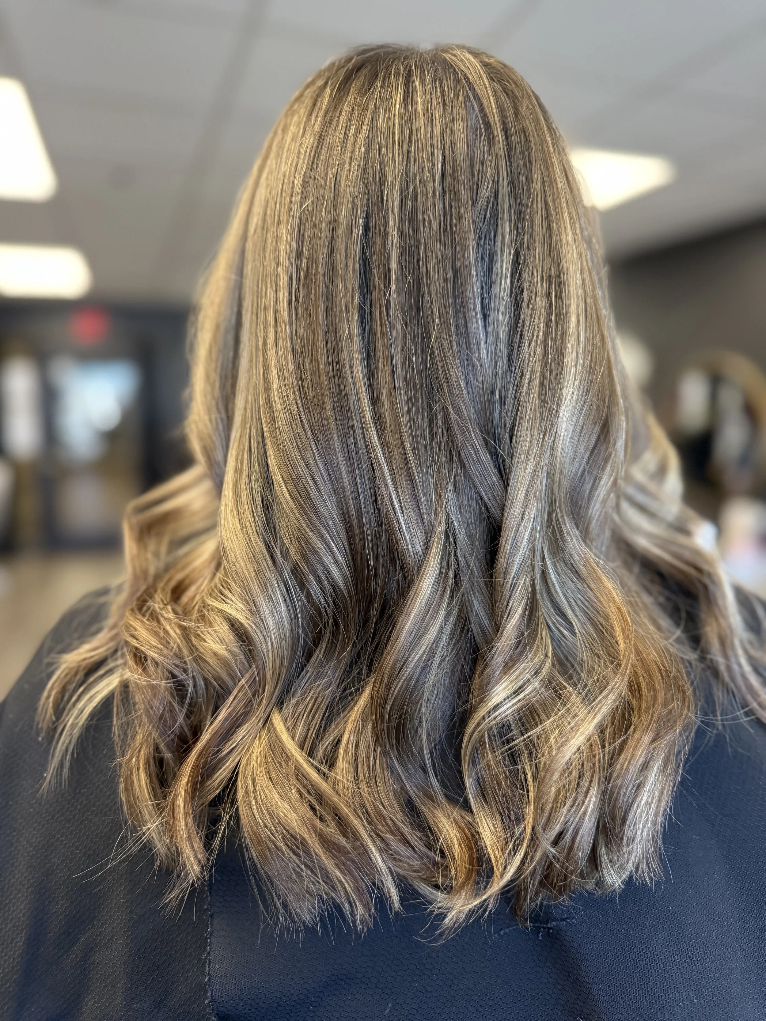 Back view of a woman with shoulder-length wavy blonde hair, wearing a black top, in a salon or similar setting.