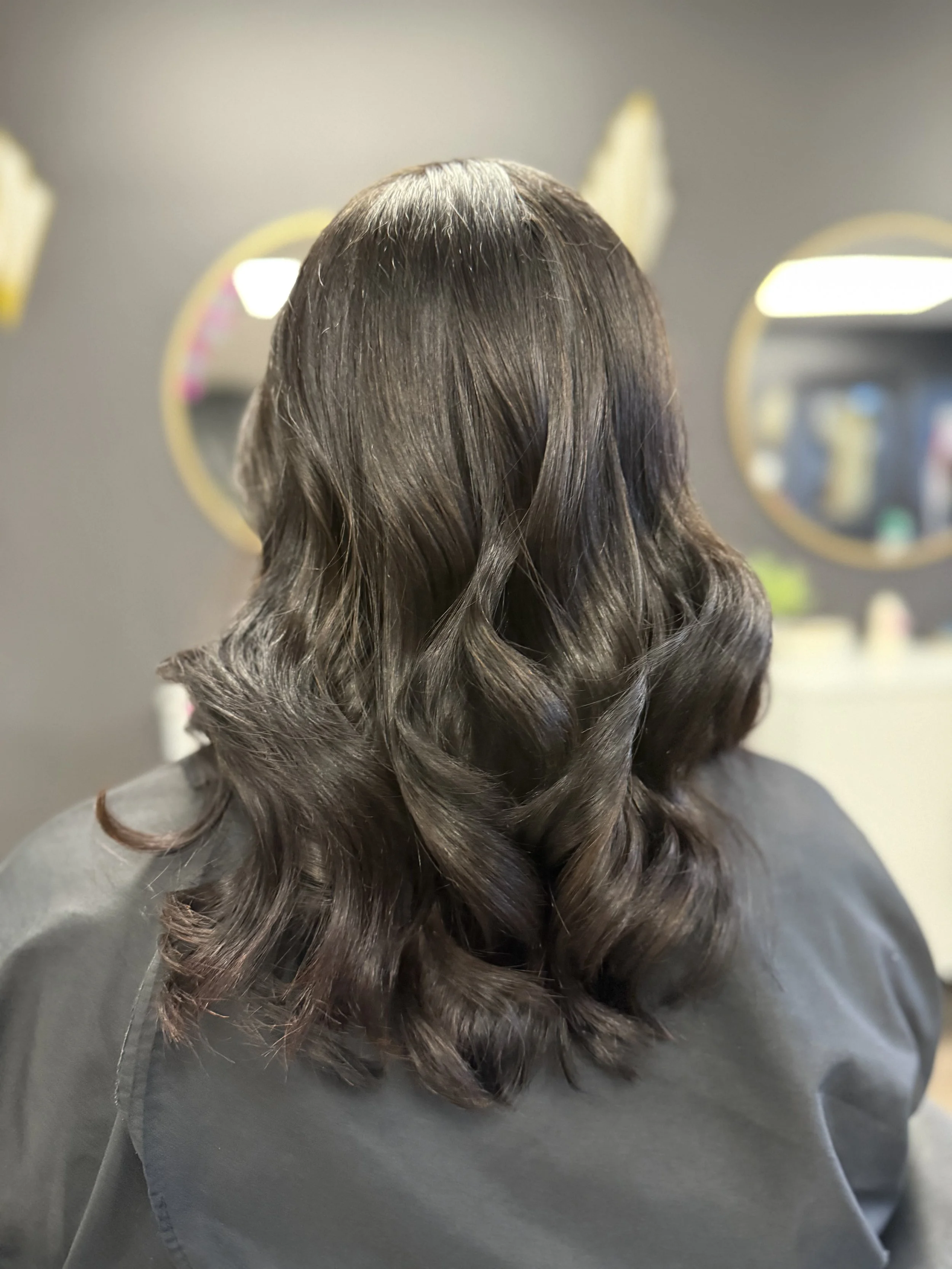 Back of a woman with shoulder-length wavy dark brown hair, sitting in a salon.