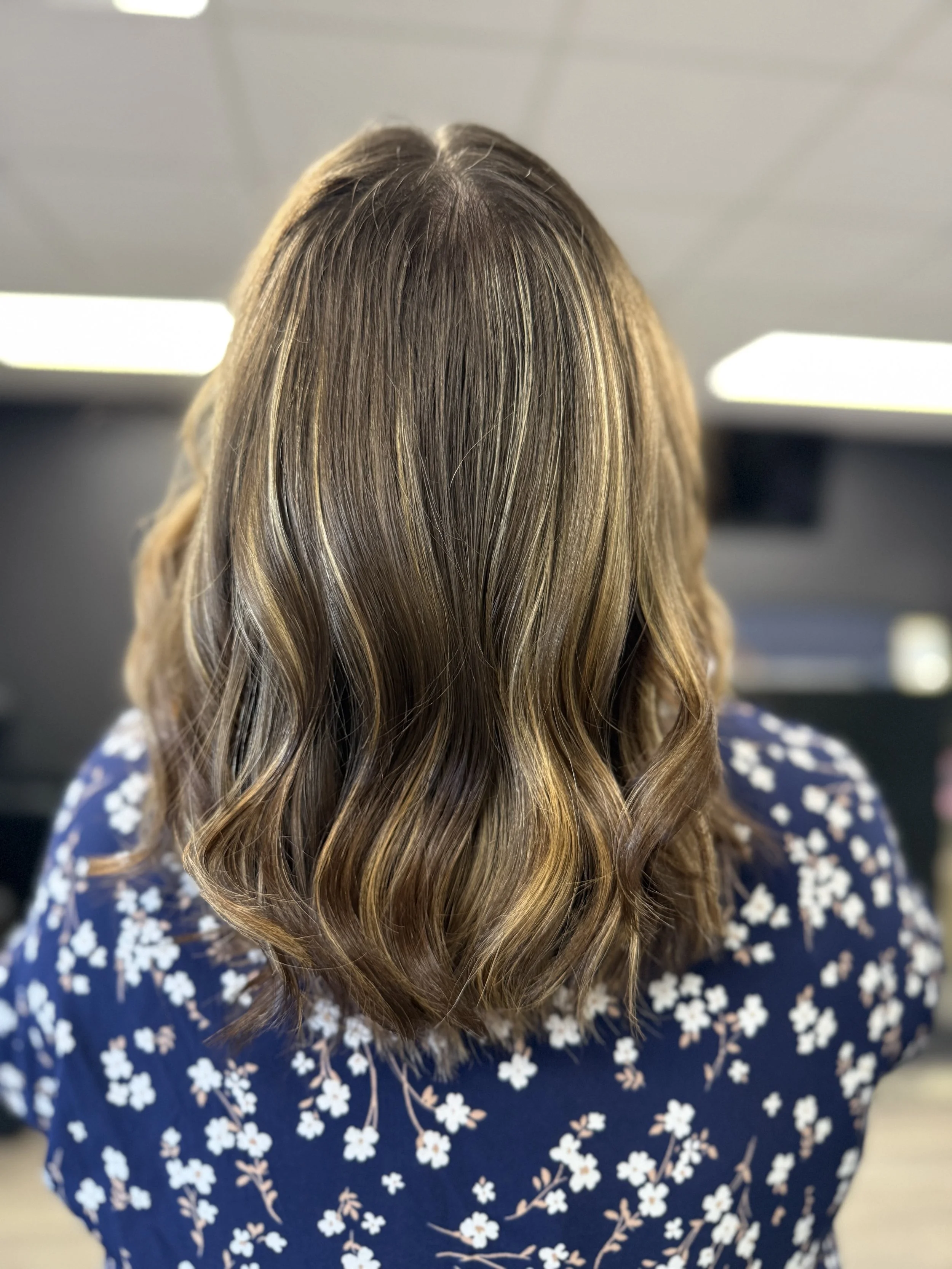 Back view of woman with medium-length wavy brown hair with blonde highlights, wearing a navy blue shirt with white floral pattern, in an indoor setting.
