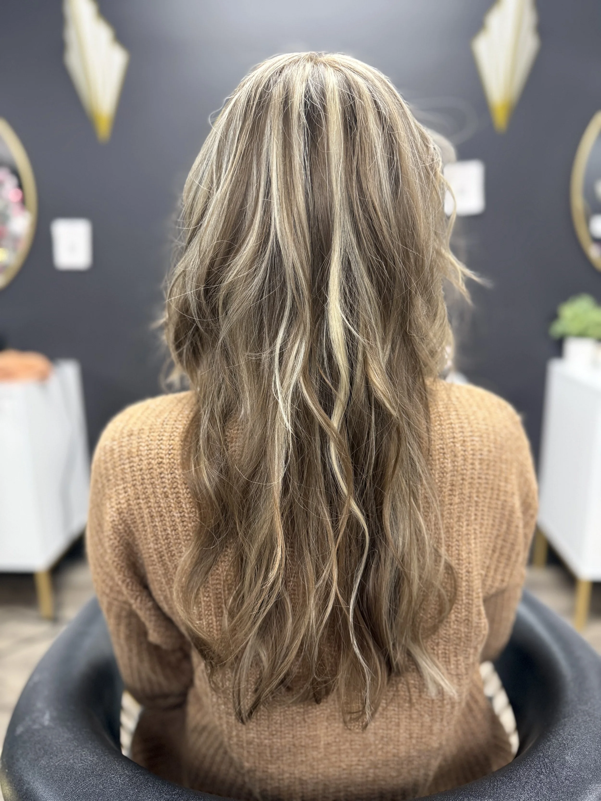 Back view of a woman with long, wavy, blonde hair sitting in a salon chair, wearing a brown sweater.