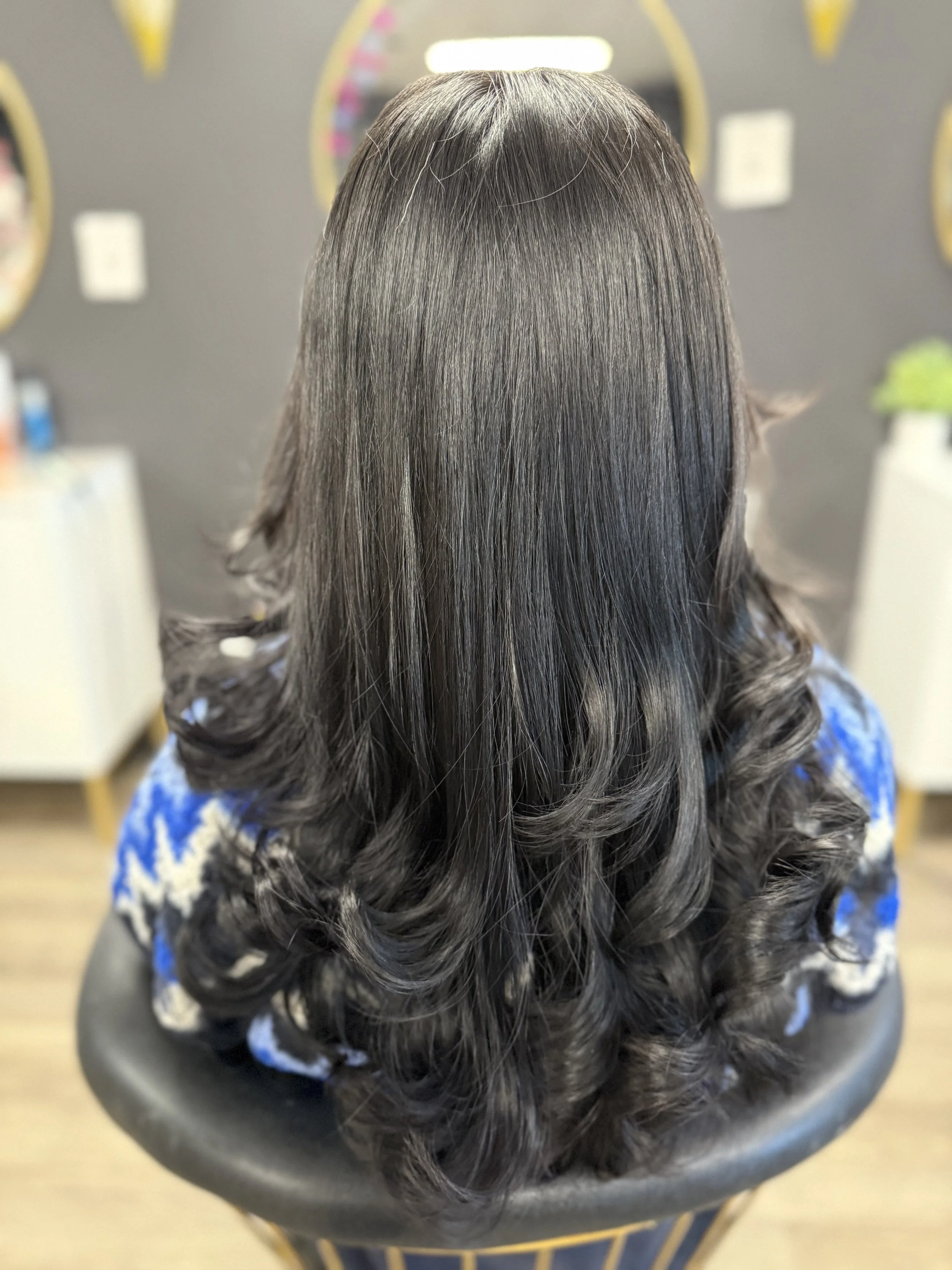 Back of a woman's head with shiny, dark, wavy hair, seated in a salon chair.
