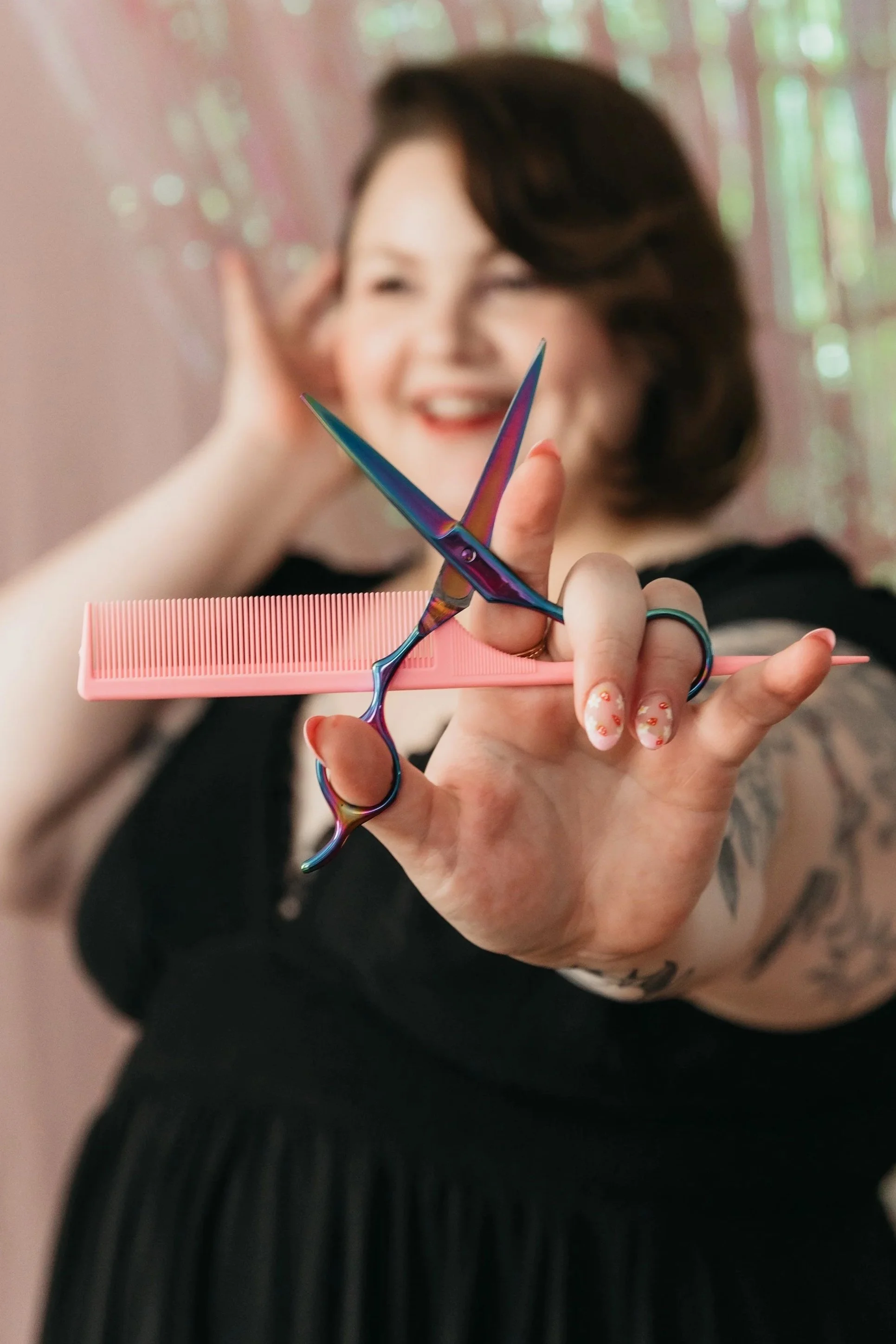 A person holding a pair of rainbow-colored scissors balanced on a pink comb with a salon or hairdressing setting in the background.