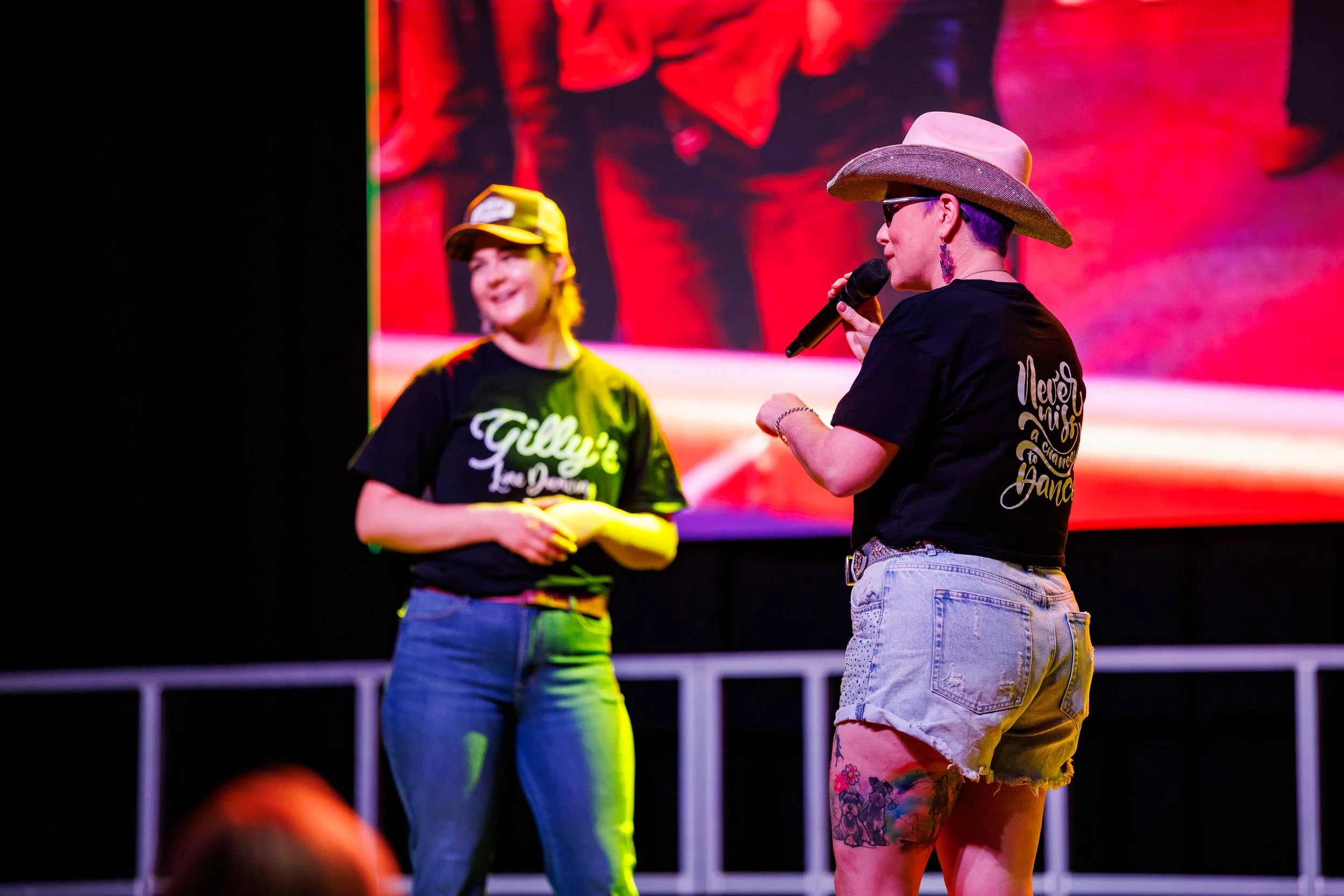 Two women on stage, one speaking into a microphone. Both are wearing hats and casual clothing. The woman in the foreground has a large hat, sunglasses, and earrings, with tattooed legs. The background features bright, colorful lighting and a large screen display.