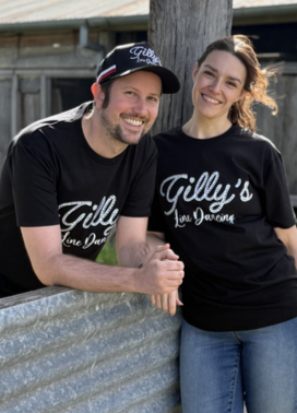 A man and woman smiling outdoors, leaning on a metal railing, both wearing black T-shirts with white text, and the man also wearing a black cap.