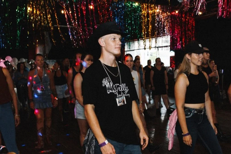 Group of young people at a dance party, some wearing cowboy hats, under colorful hanging decorations, with a dark dance floor and party lighting.
