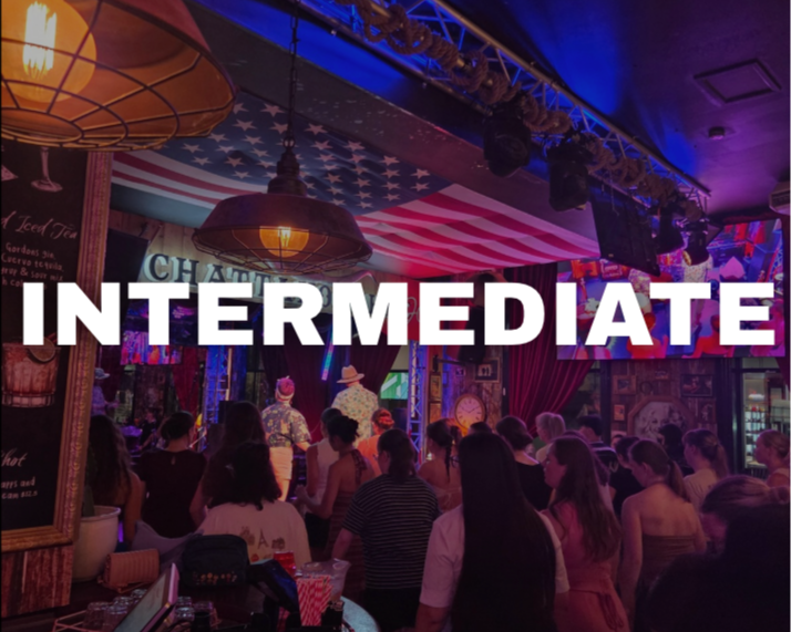 People gathered inside a bar or music venue with colorful, decorated interior and American flag ceiling, watching a performance on stage, with a sign that says 'INTERMEDIATE' in front.