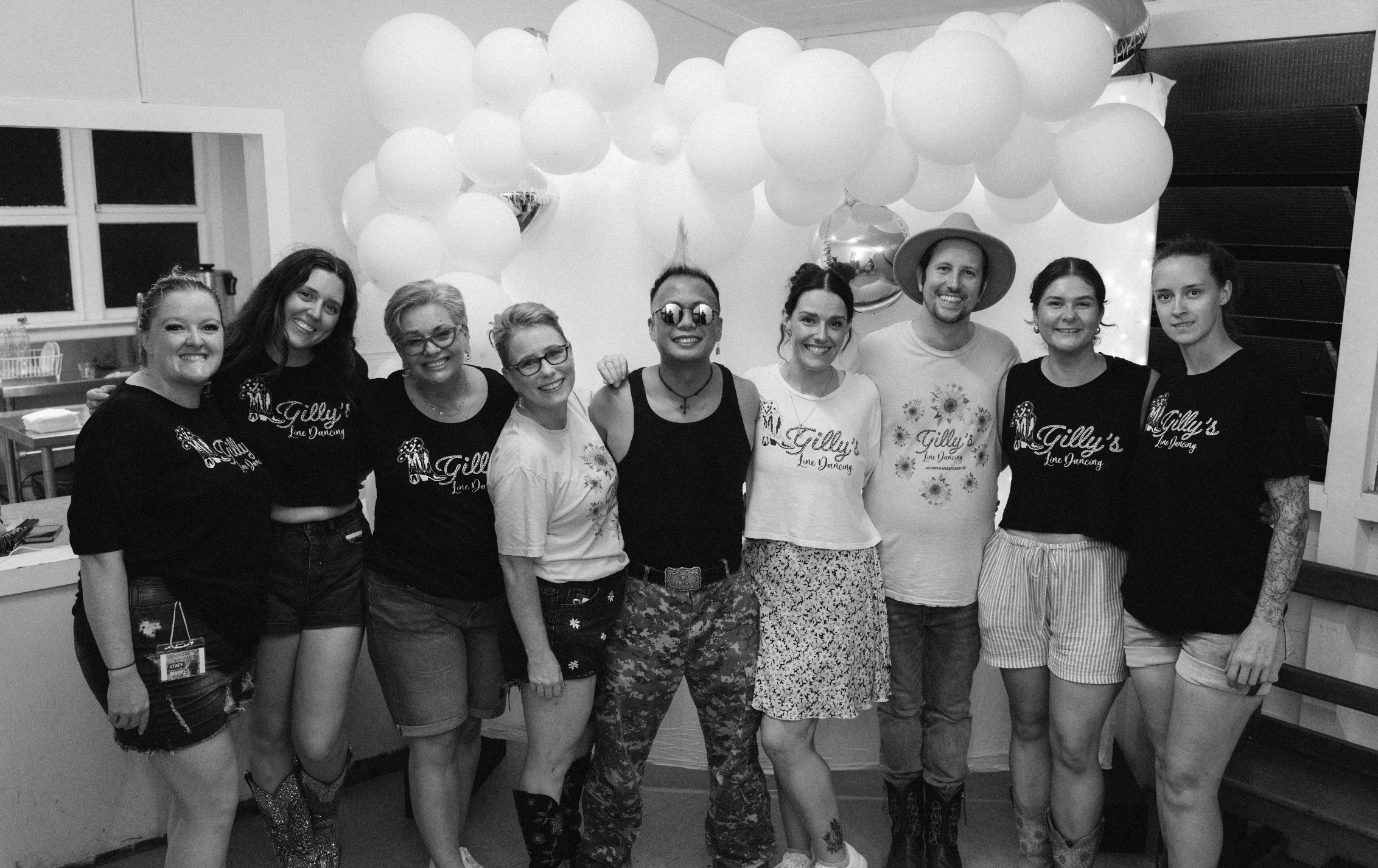 A group of nine women and one man standing indoors in front of a wall decorated with balloons, smiling at the camera. Some women are wearing shirts with 'Gilly's Line Dancing' printed on them.