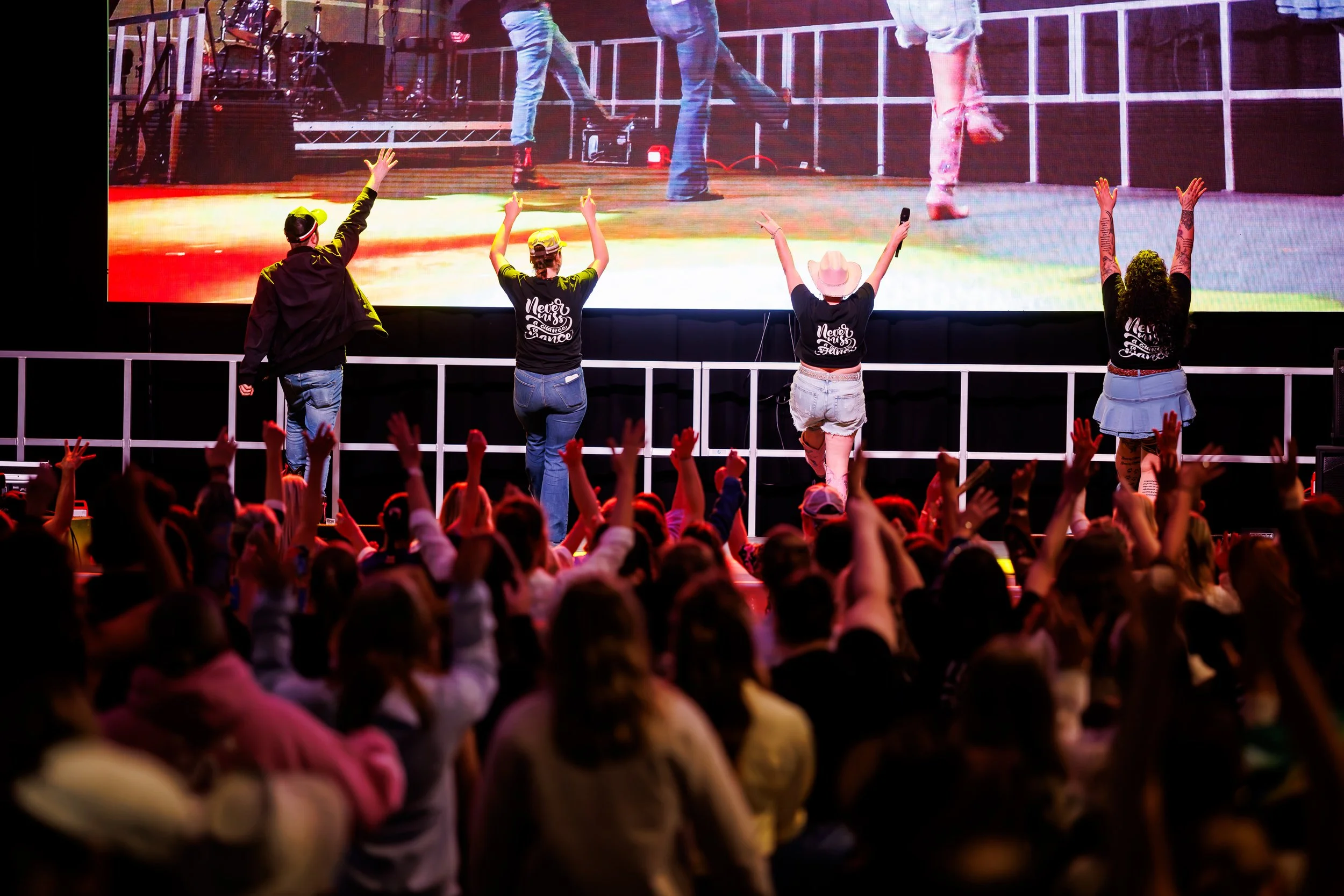 People on stage performing at a concert with an audience in front, some with arms raised. The stage has a large screen behind showing close-up views of the performers.