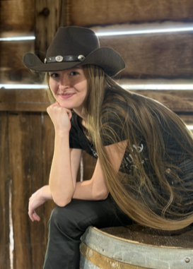 Young woman wearing a cowboy hat, sitting on a wooden barrel inside a rustic wooden building, smiling at the camera.
