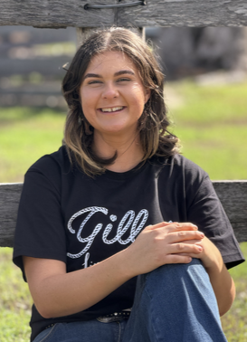 Young girl with brown hair smiling, wearing a black T-shirt and blue jeans, sitting outdoors on a sunny day.
