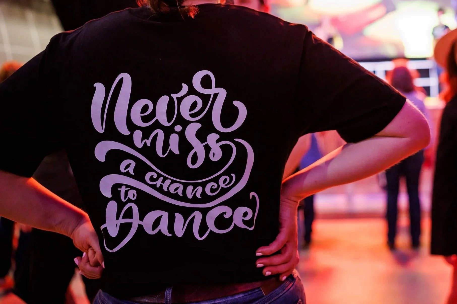Person wearing a black T-shirt with the text "Never miss a chance to dance" written in large purple letters, standing with hands on hips in a dimly lit dance floor or party setting with colorful lighting and other people in the background.