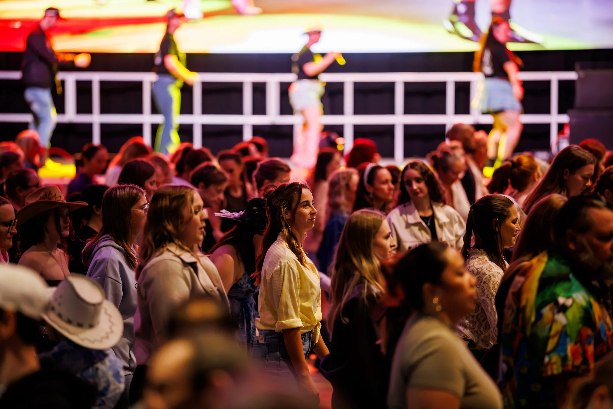 Crowd of people watching a stage with four performers dressed in cowboy outfits, including hats and boots, with a bright screen in the background.
