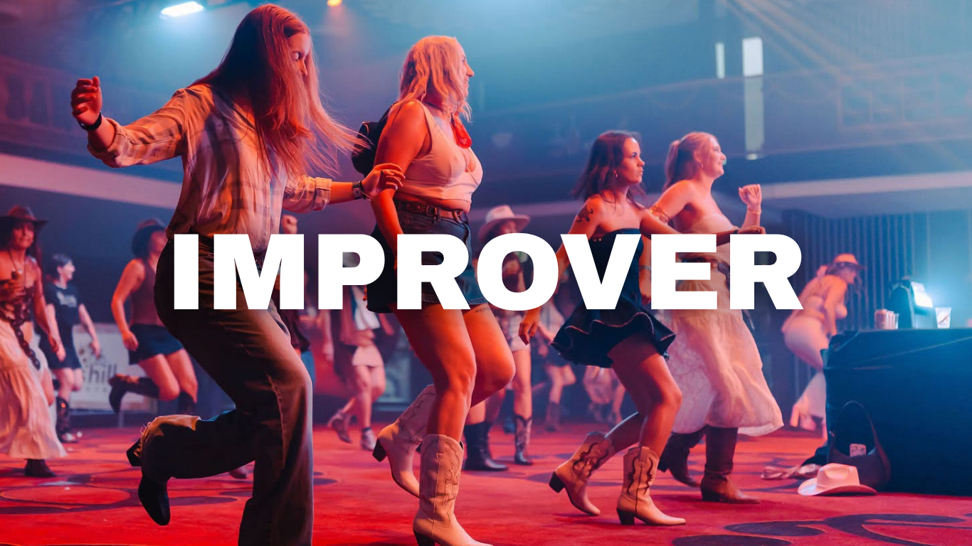 Women dancing at a country music concert or line dancing event, wearing Western-style clothing, cowboy boots, and hats, inside a large concert venue with stage lights and audience in the background.