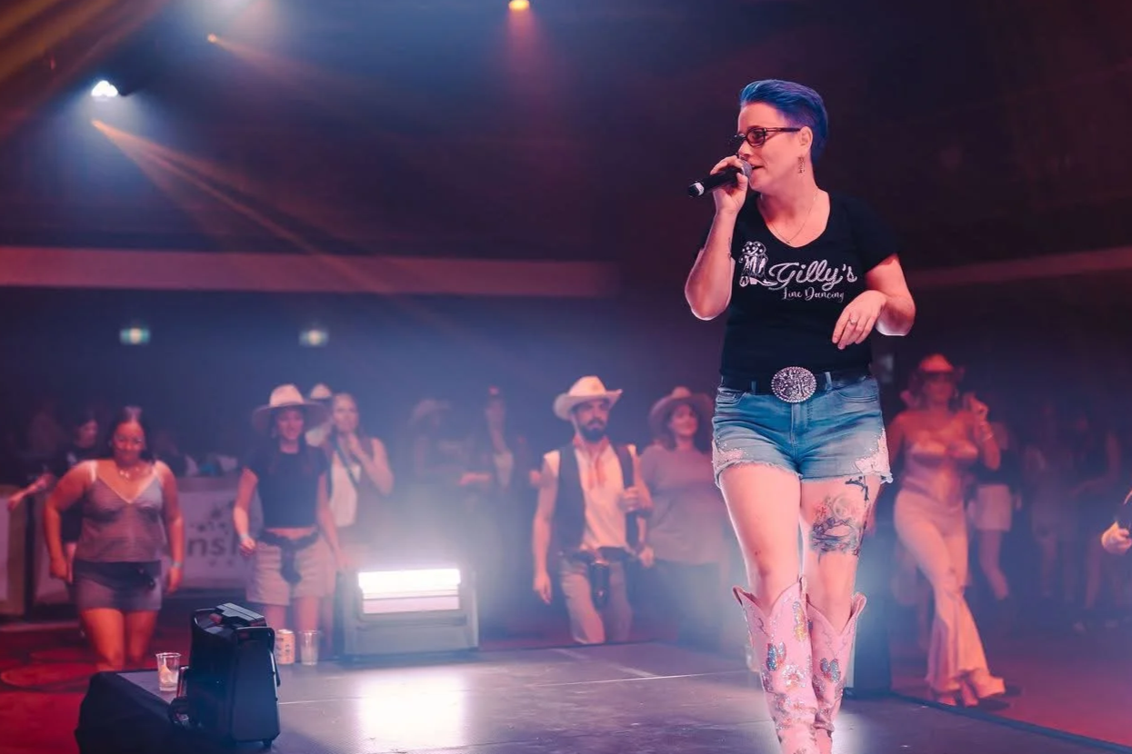 A woman with short blue hair, glasses, and tattoos is singing into a microphone on stage at a country-themed event, wearing a black T-shirt, denim shorts, and pink cowboy boots. People in cowboy hats and casual attire are dancing and watching in the background.