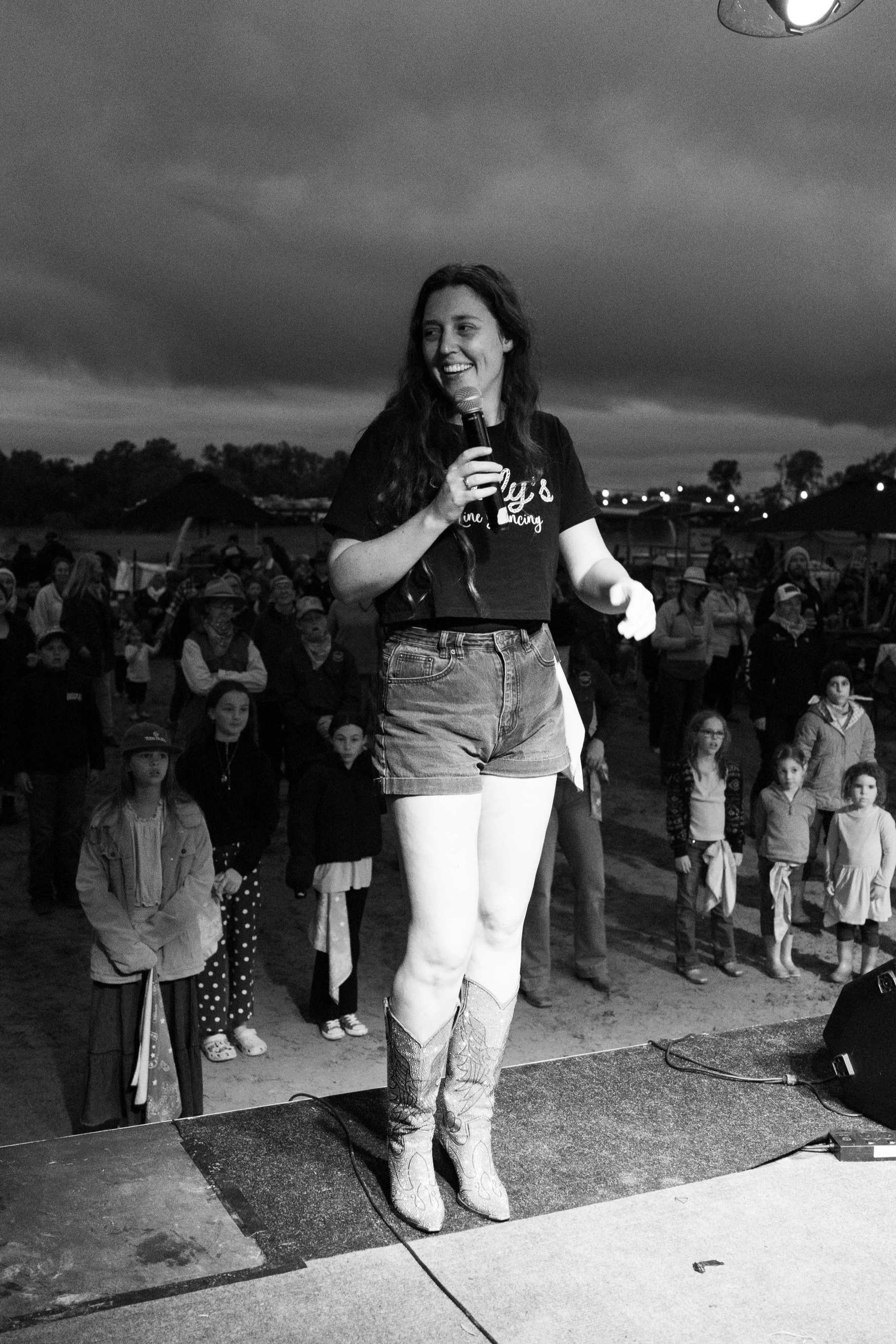 A woman standing on stage with a microphone, wearing cowboy boots and shorts, smiling while addressing a crowd of children and adults outdoors during evening or night.