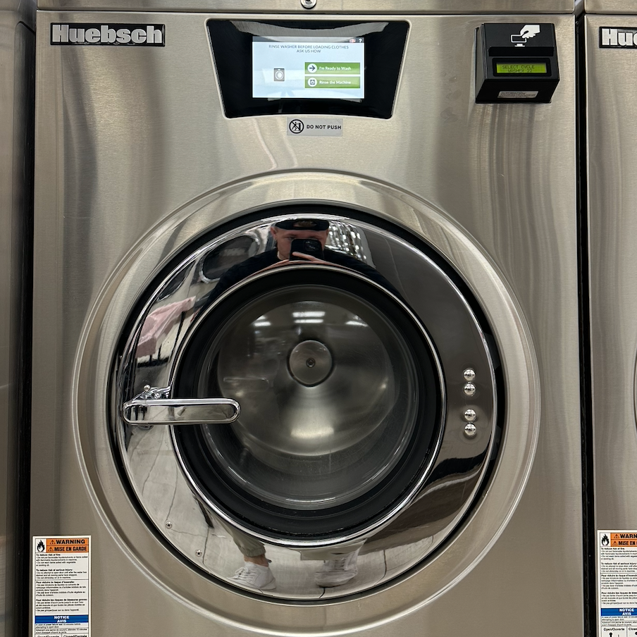 Front view of a stainless steel Huebsch commercial washing machine with a digital display and control panel at the top, and a reflective door showing the person taking the photo in a laundry room.